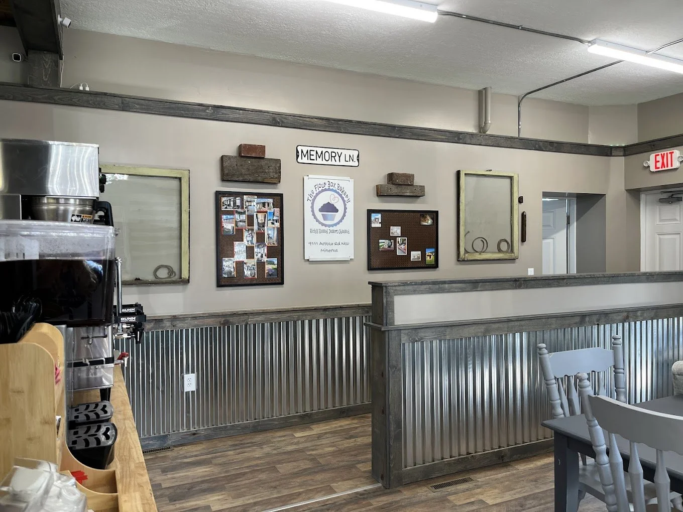 Interior of a bakery or cafe with a reception counter, wall decor including photos, signs, and small shelves, and a dining area with wooden chairs and tables.