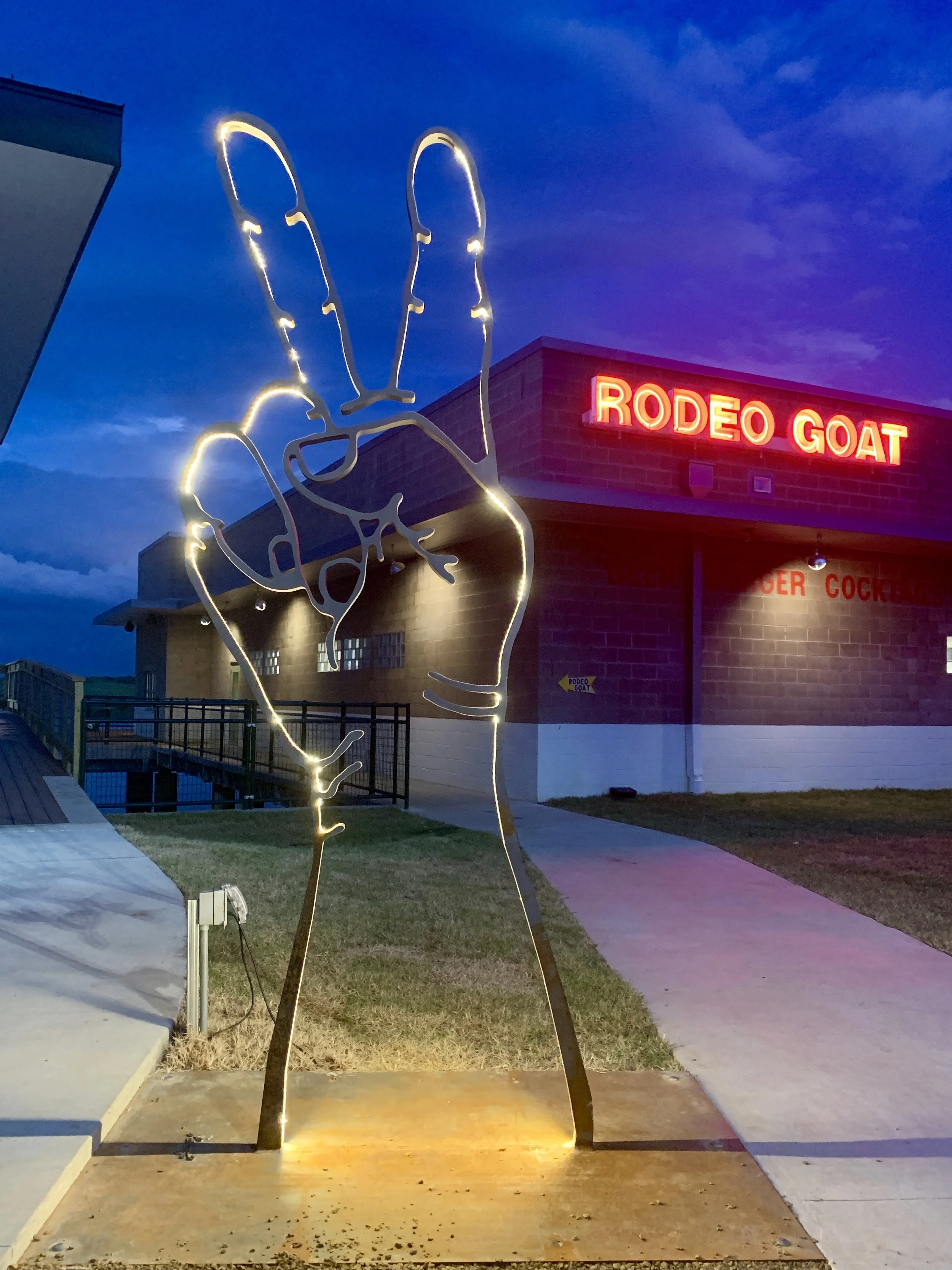 LED-lit sculpture of a 10Ft tall steel hand making a peace sign, outside of 'RODEO GOAT' at Cypress Waters at dusk.