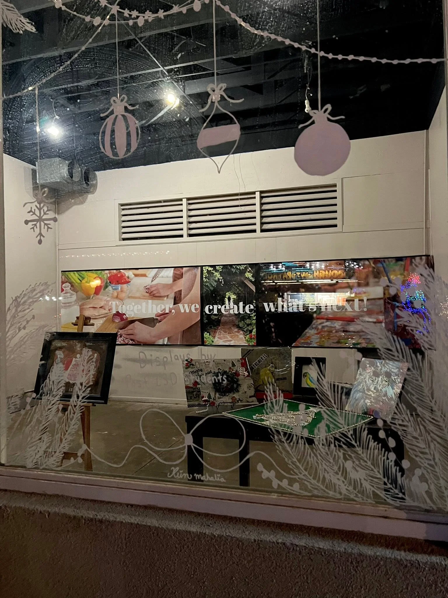 Display window with Christmas decorations and festive signage, featuring ornaments, snowflakes, and holiday-themed items at a shopping mall.