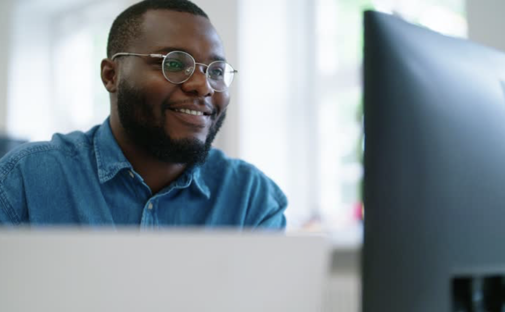 A man with glasses and a beard smiling while working on a computer.