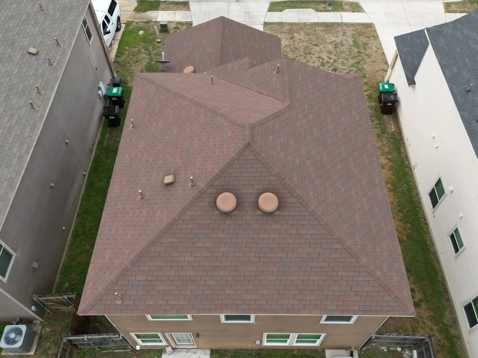 Aerial view of a house with a brown shingle roof, flanked by neighboring houses, with driveways, trash bins, and grassy areas visible.