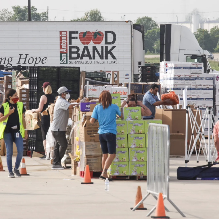 People working at a food bank distribution site with boxes of supplies and a food bank truck in the background.