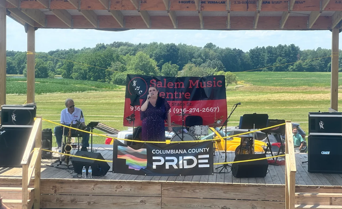 A woman singing into a microphone on an outdoor stage with a man playing a guitar or keyboard to her left and a drummer behind her. The stage has a banner reading 'Salem Music Centre' and another banner with 'Columbiana County Pride.' A field with trees is visible in the background.