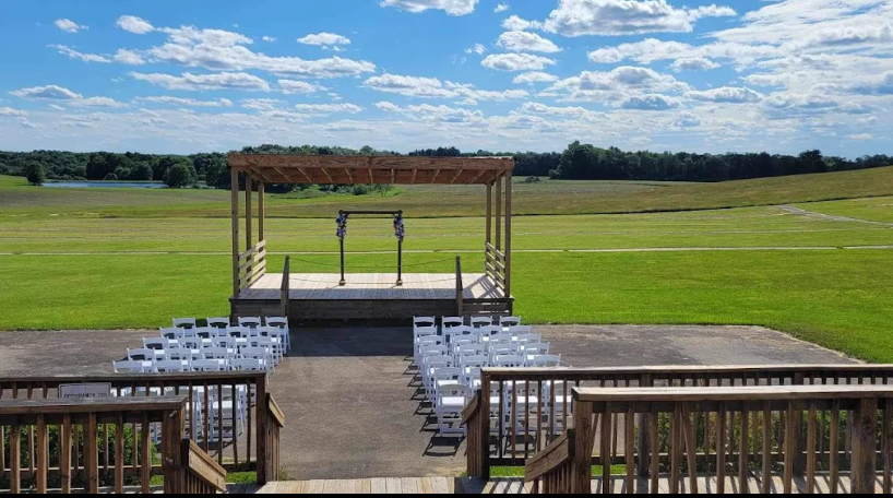An outdoor event venue with a wooden stage and white chairs set up on a grassy field beneath a partly cloudy sky.