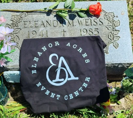 Gravestone for Eleanor M. Weiss (1941-1985) with a black cloth displaying the Eleanor Acre's Event Center logo, surrounded by flowers.