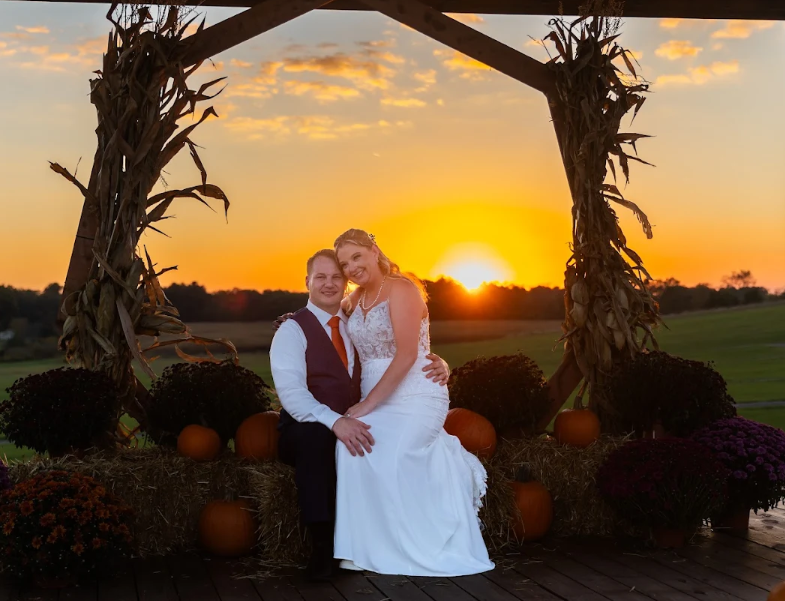 A couple dressed in wedding attire sitting together on a hay bale, with the sunset in the background. The bride is in a white gown, and the groom is in a white shirt with a vest and tie. The scene features autumn pumpkins and harvest decorations, with corn stalks framing the couple and a field in the distance.