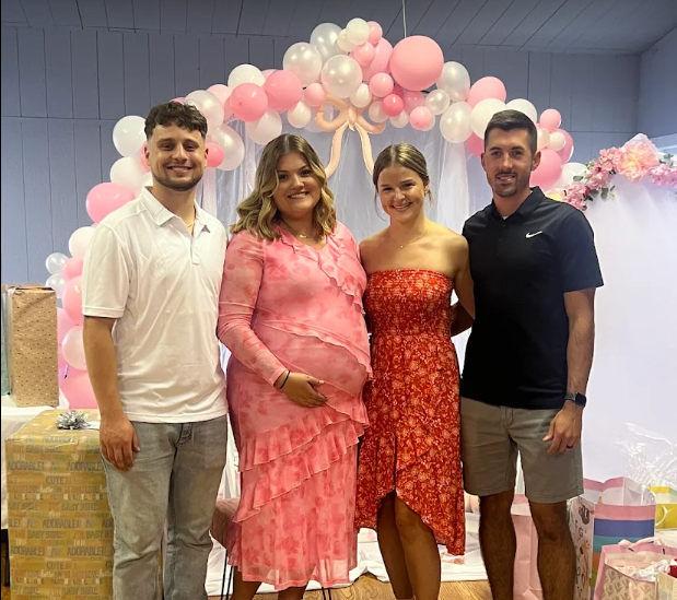 Four young adults, two men and two women, standing together at a celebration or party with pink and white balloons and decorations in the background. The woman in the middle is pregnant and wearing a pink dress, while the others are dressed casually.