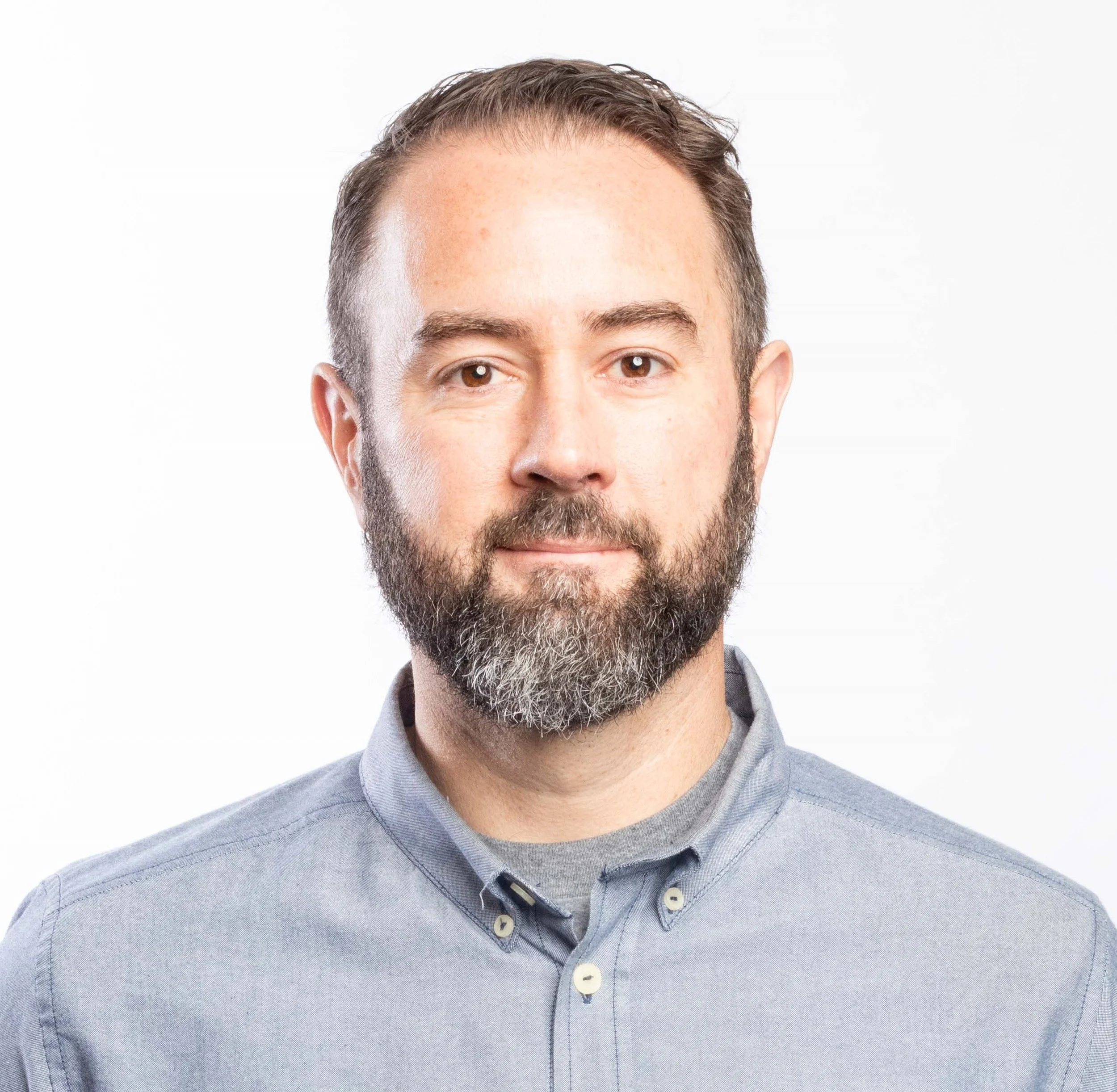 Close-up of a man with a beard wearing a light blue button-up shirt, smiling slightly, against a white background.