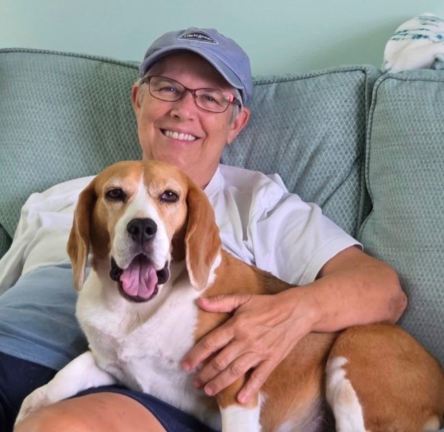 A smiling woman with glasses and a blue cap sitting on a light green couch, holding a happy beagle dog with its tongue out.