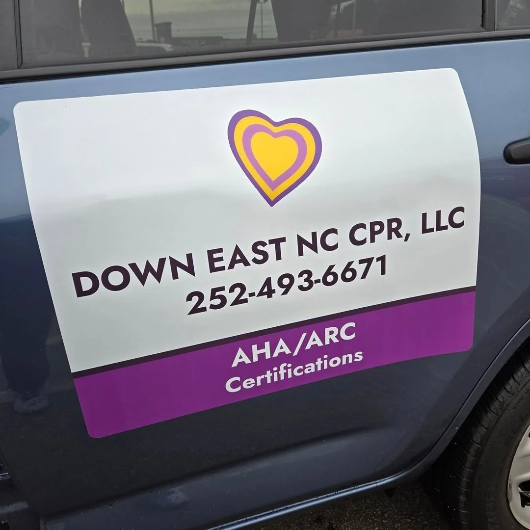 Sign on car door for Down East NC CPR LLC with purple and yellow heart logo, phone number 252-493-6671, and text AHA/ARC Certifications.