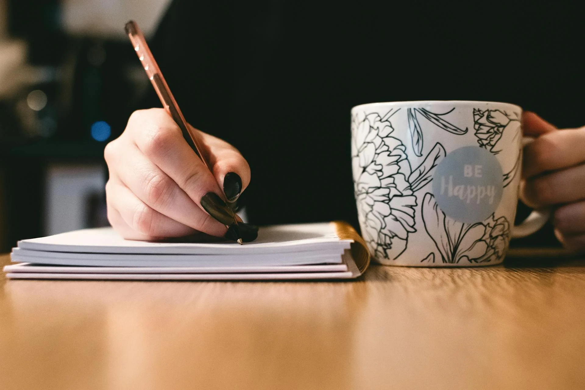 A person's hand with black painted nails writing in a notebook with a rose gold pen, next to a mug with floral design and the words 'Be Happy' on a wooden surface.