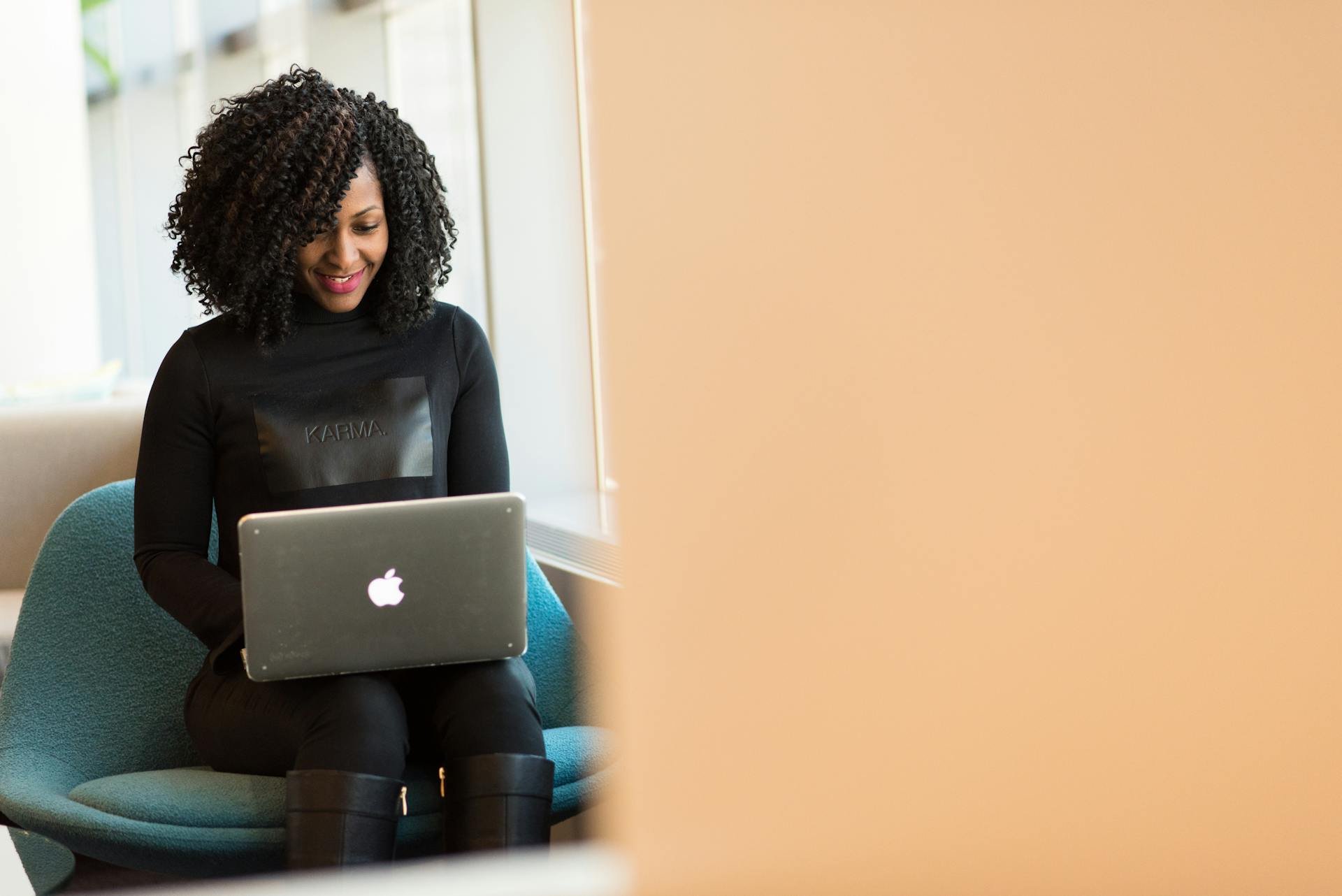 A woman with curly hair, wearing a black sweater, sitting on a light blue chair, looking at an Apple MacBook laptop, in a bright and modern indoor space.