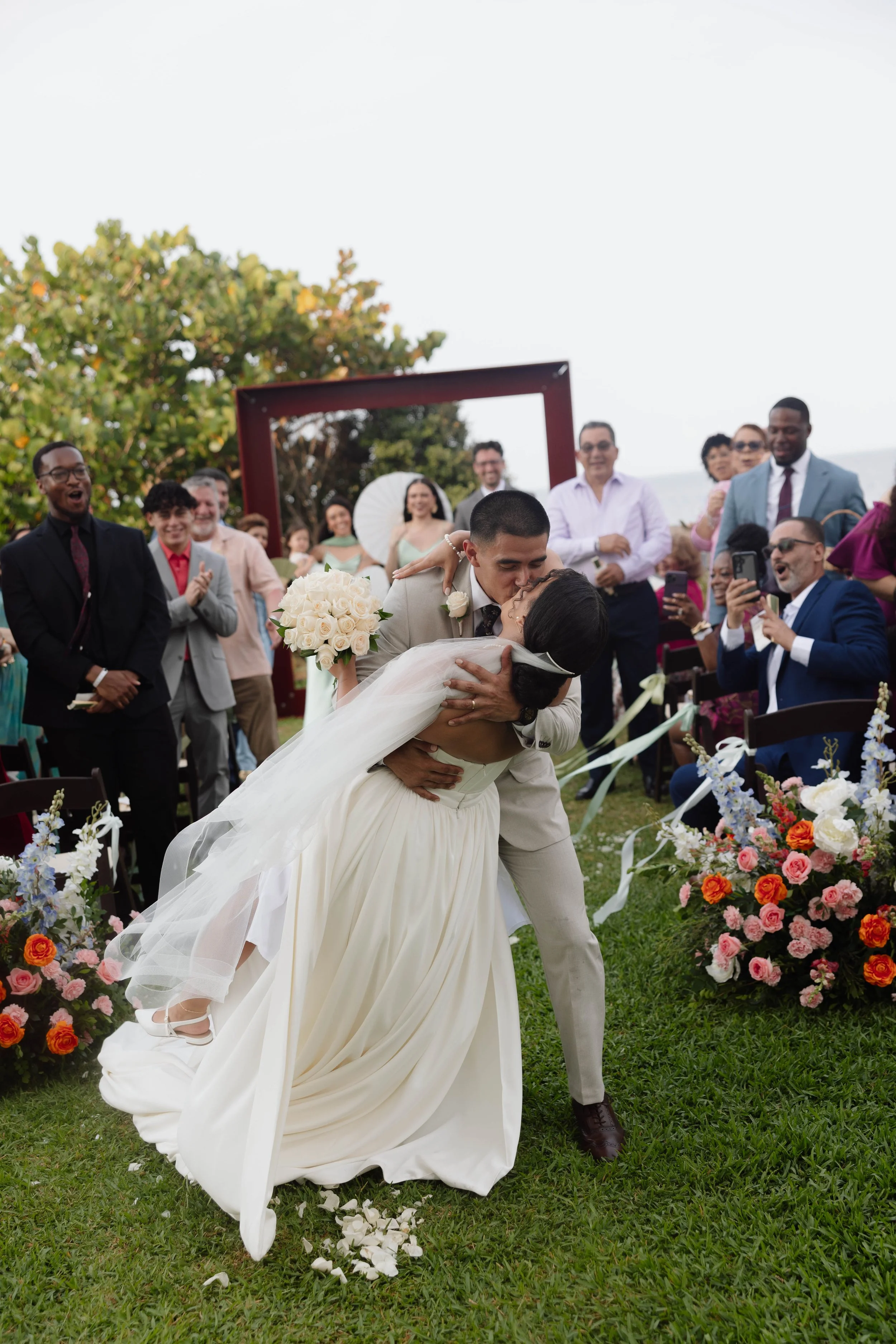 A newlywed couple sharing a kiss while dancing at an outdoor wedding ceremony, with guests clapping and celebrating around them, colorful flowers, and lush greenery in the background.