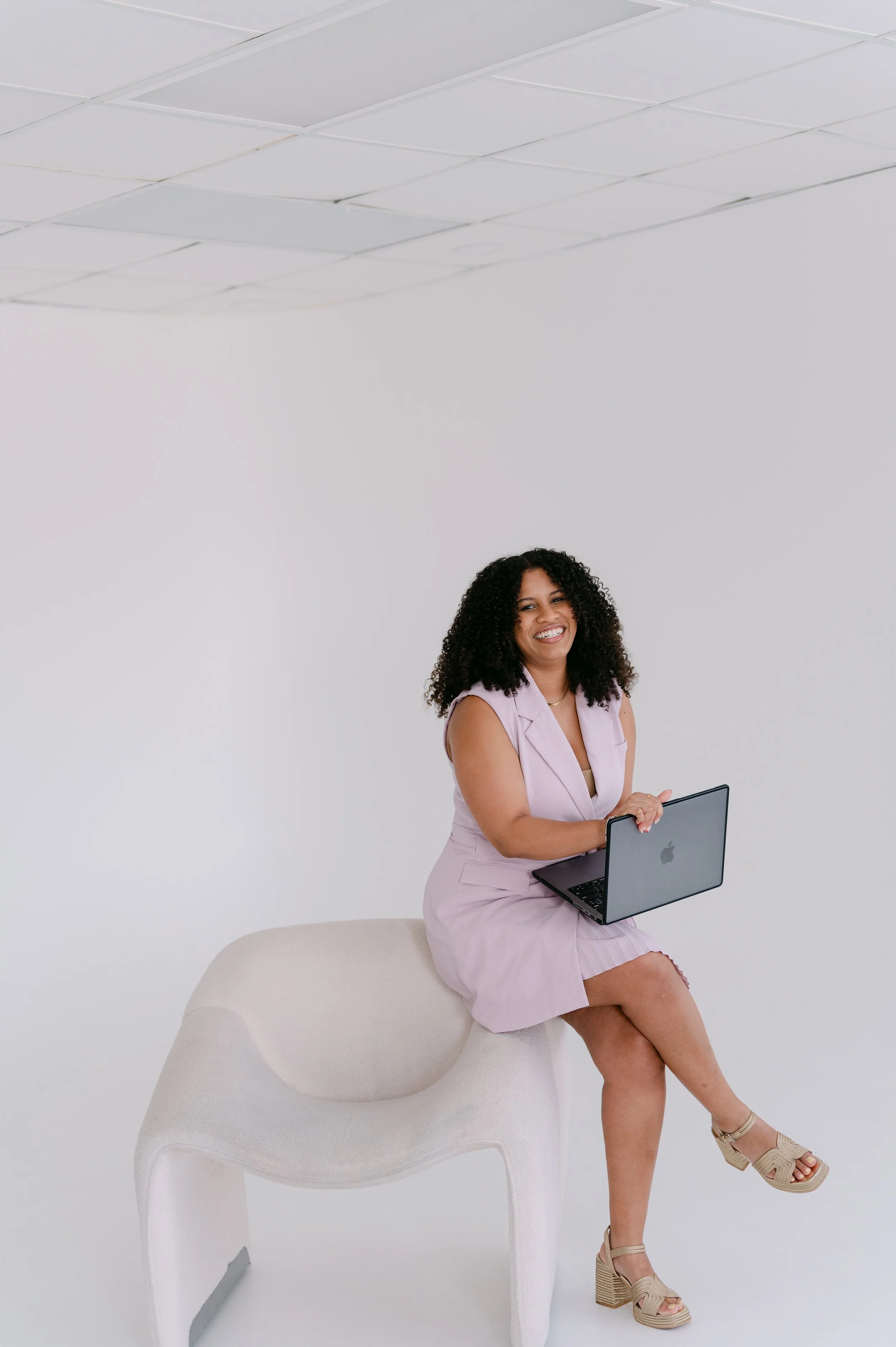 A woman with curly hair sitting on a white curved chair, smiling and holding an open laptop on her lap. She is wearing a light pink sleeveless dress and tan platform sandals. The background is plain white with a ceiling grid visible above.