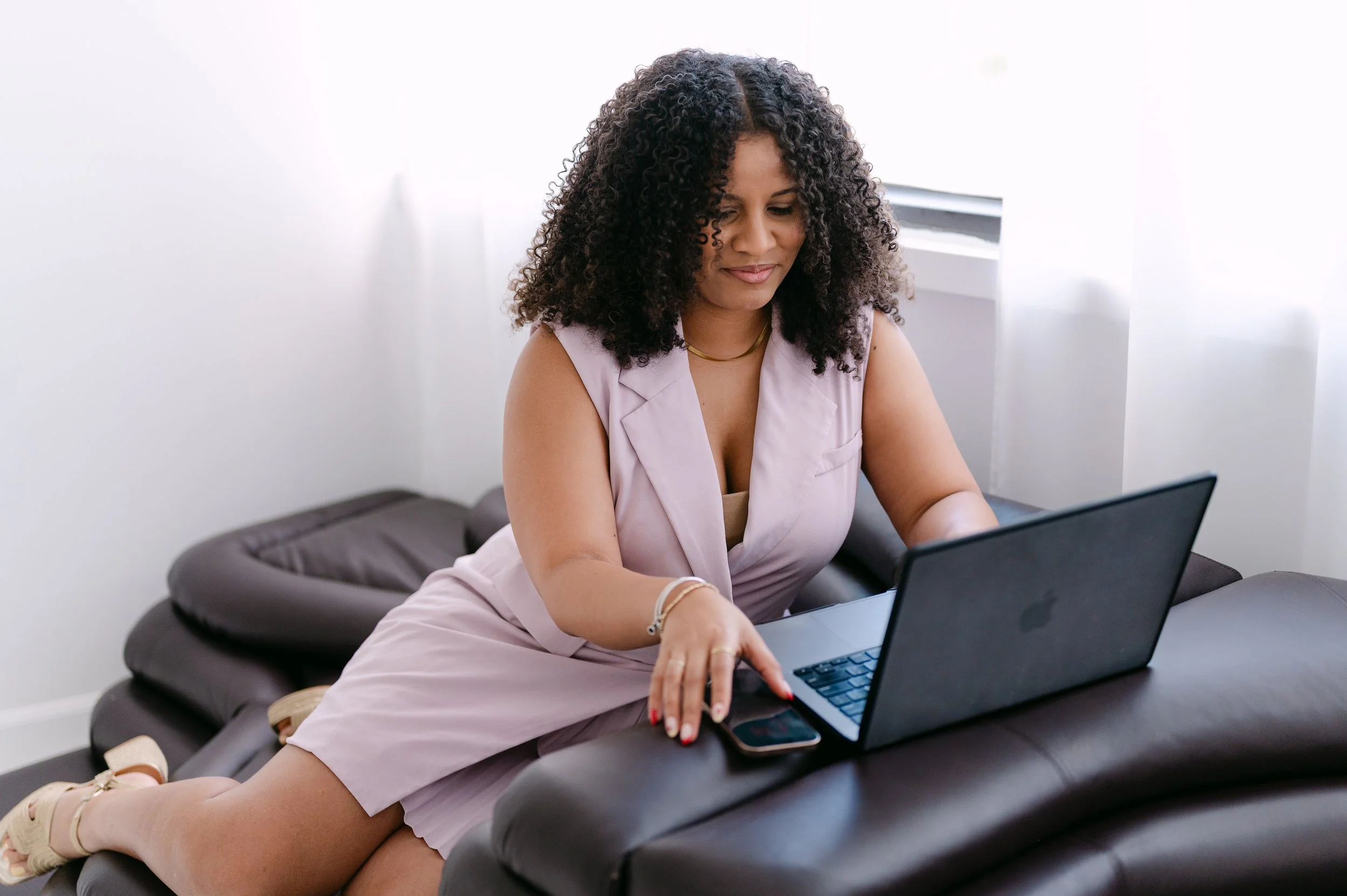 A woman sitting on a black leather sofa using a laptop computer with a phone next to her. She is wearing a light purple sleeveless blouse, beige high-heeled sandals, and gold jewelry. She has curly dark hair and a light skin tone, with a serene expression. The room is bright with white walls and a window.
