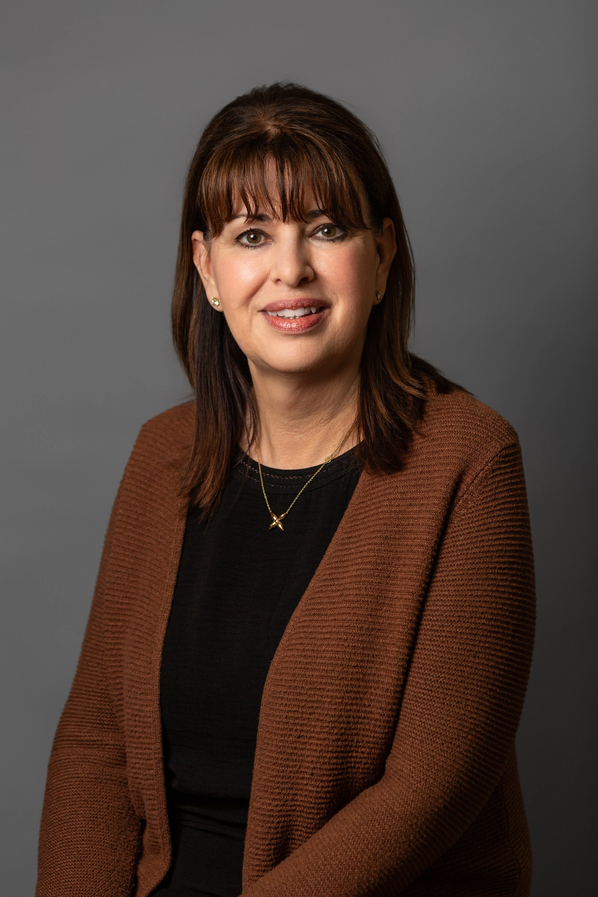 A woman with dark brown hair, wearing a black top, a brown blazer, and gold jewelry, smiling at the camera against a grey background.