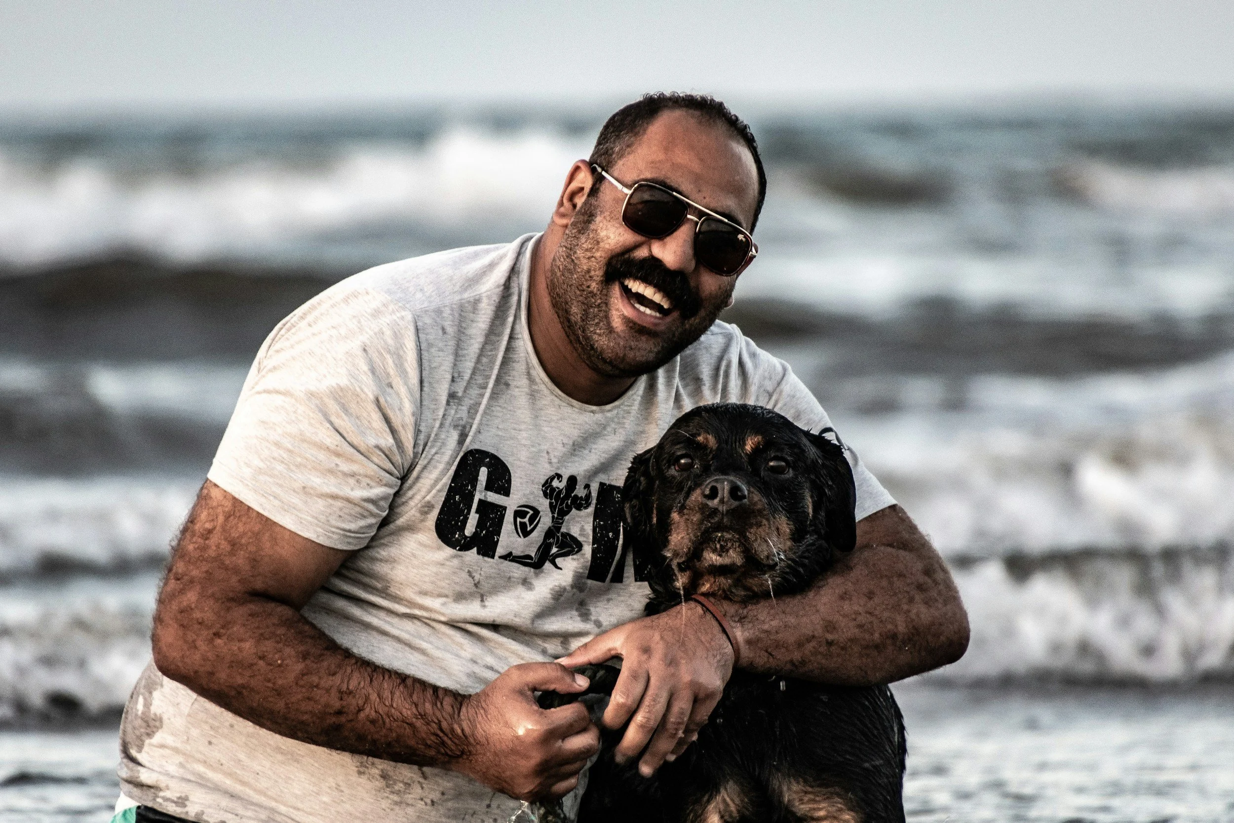 A man with sunglasses and a beard wearing a gray T-shirt holding a black and brown dog in the ocean on a beach.