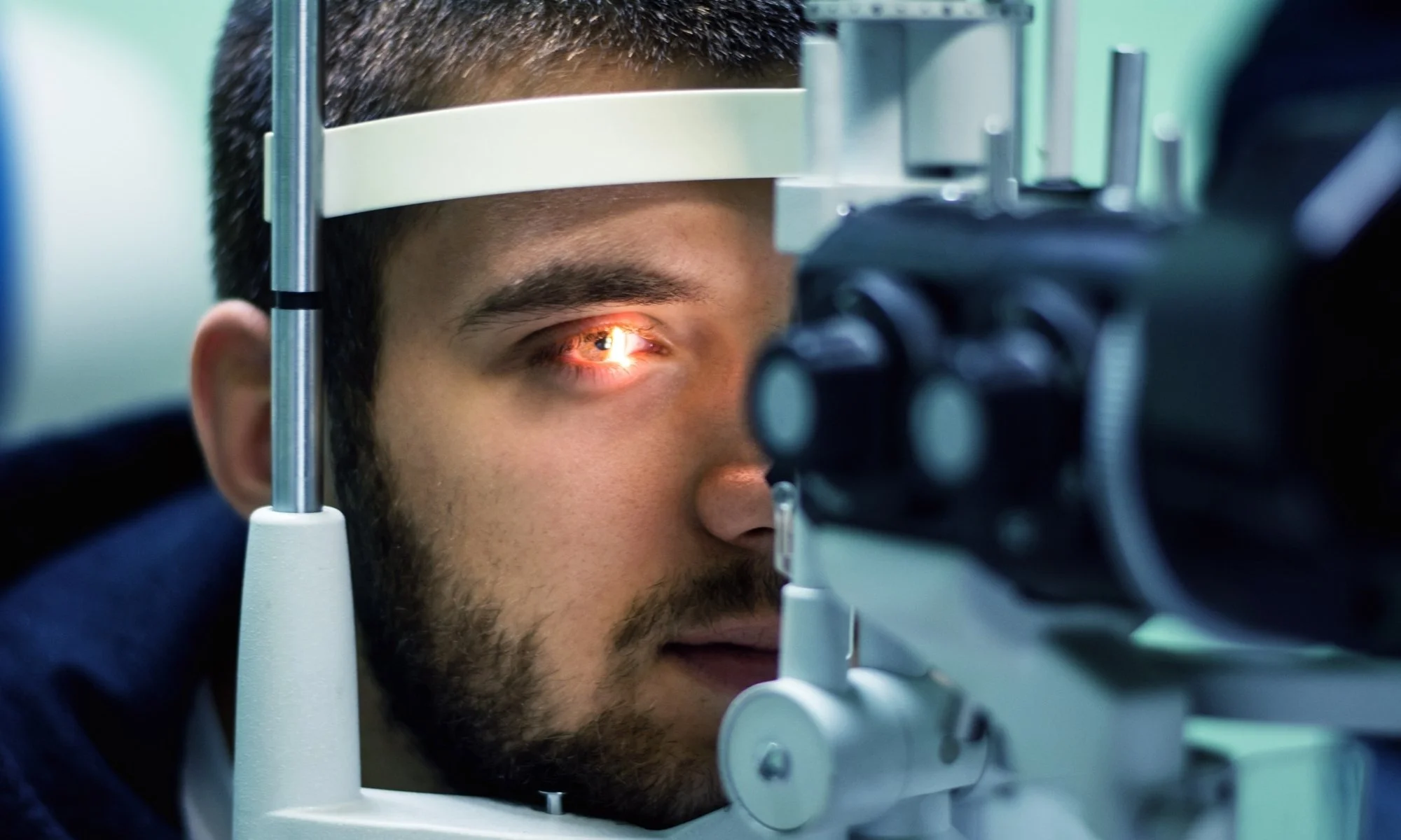 A man undergoing an eye exam using a slit lamp, with one eye positioned close to the equipment and a reflection of a bright light seen in his eye.