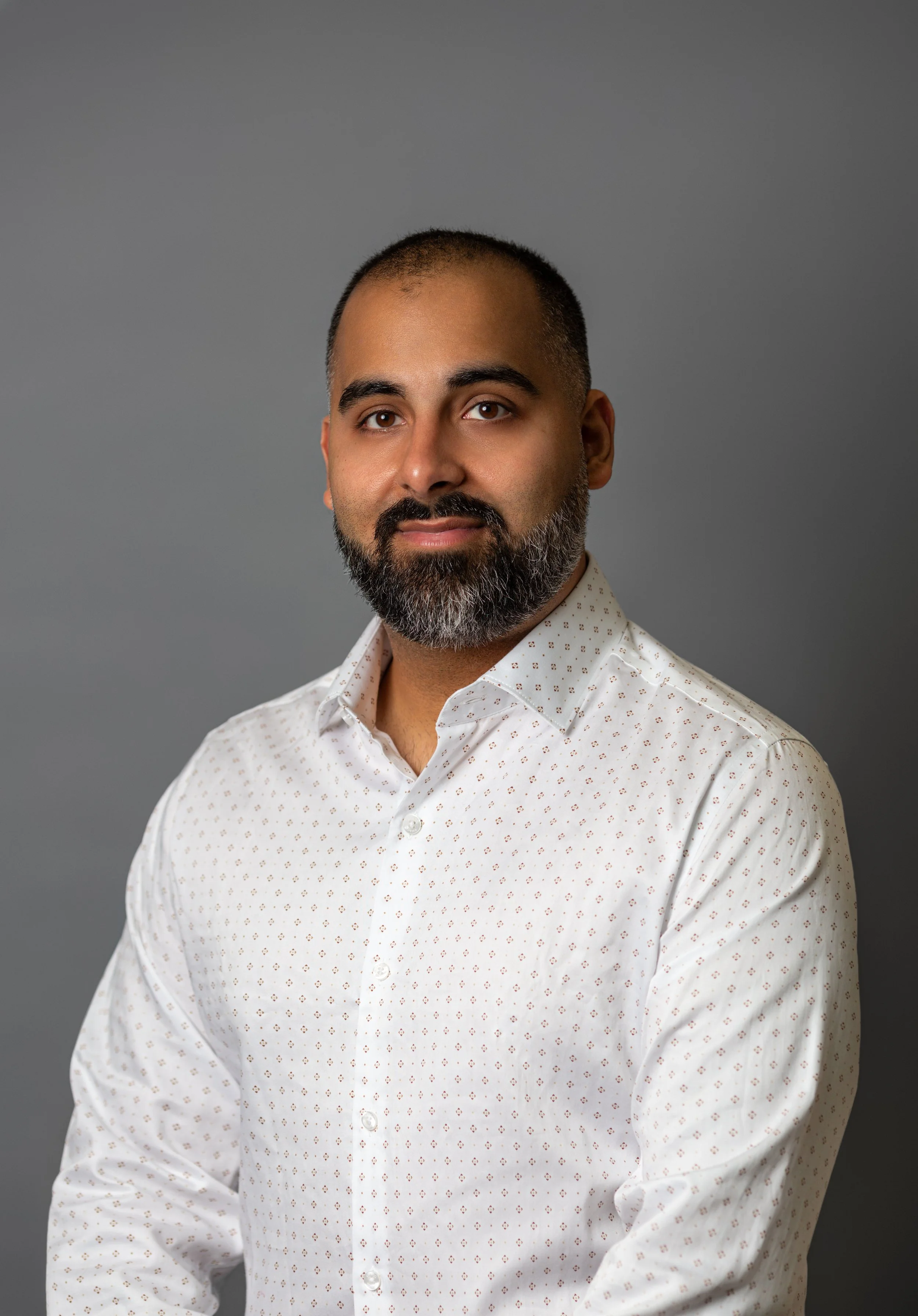A man with a beard and short hair, wearing a white button-up shirt with small pattern, standing against a gray background.
