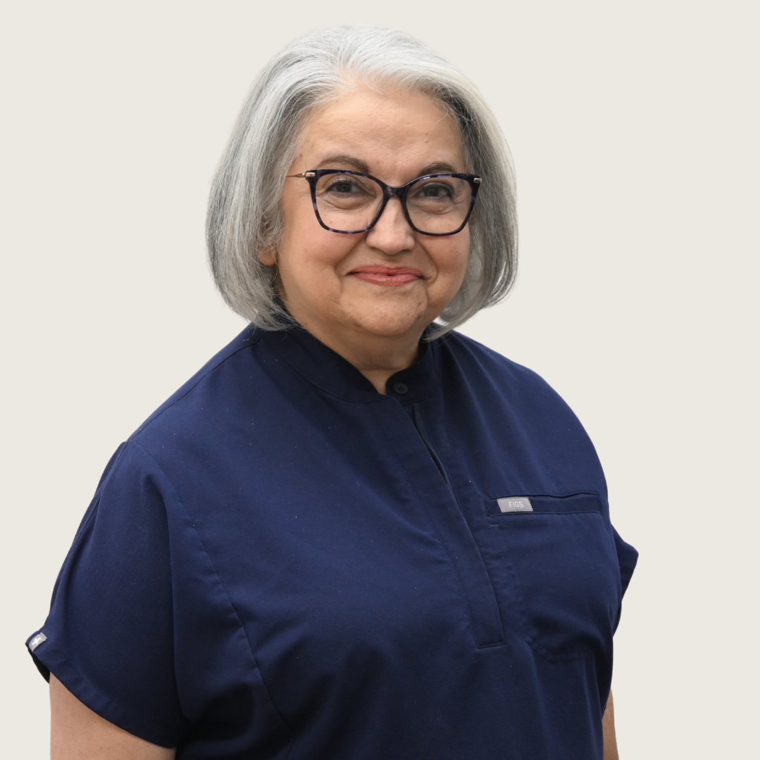 A mature woman with shoulder-length gray hair, wearing glasses and a white blouse with a bow at the neck, smiling slightly against a gray background.
