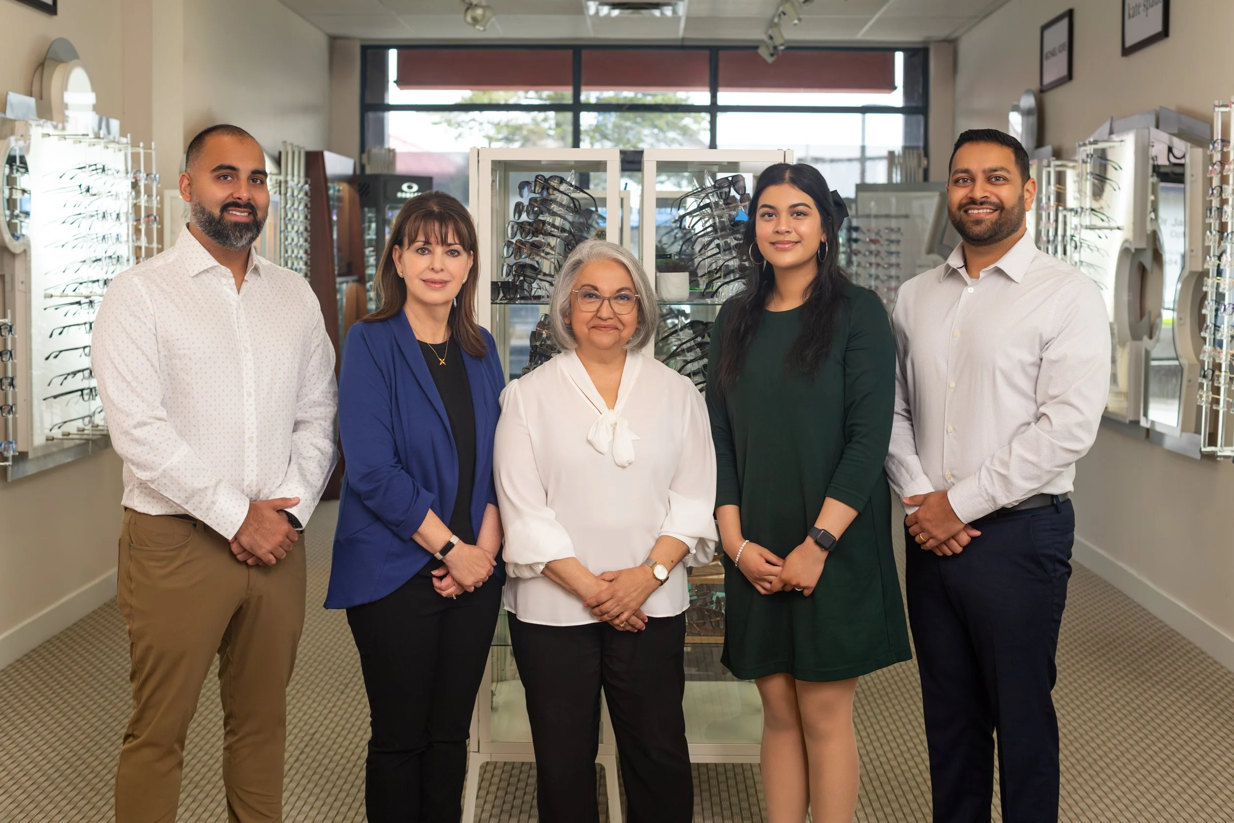 A diverse group of five people, including three women and two men, standing inside an eyewear store, smiling and posing for a photo. The background features display shelves with eyeglasses.