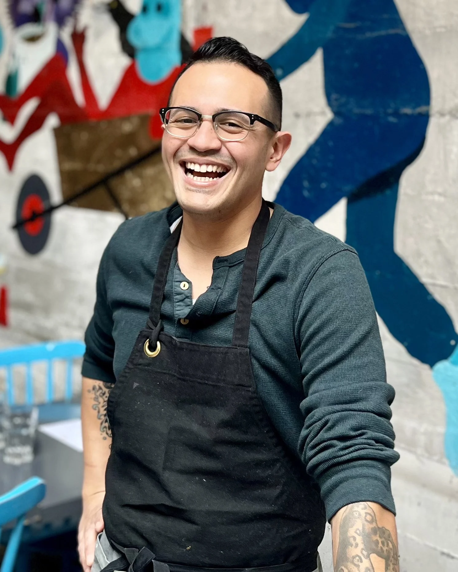 A smiling man wearing glasses and a black apron, standing in front of a colorful graffiti wall.