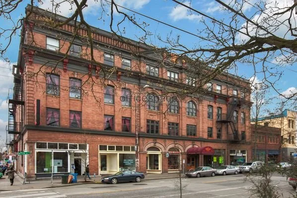 A multi-story brick building with commercial storefronts on the ground level, situated along a city street with parked cars and pedestrians. The building has decorative arched windows and fire escapes on the exterior.