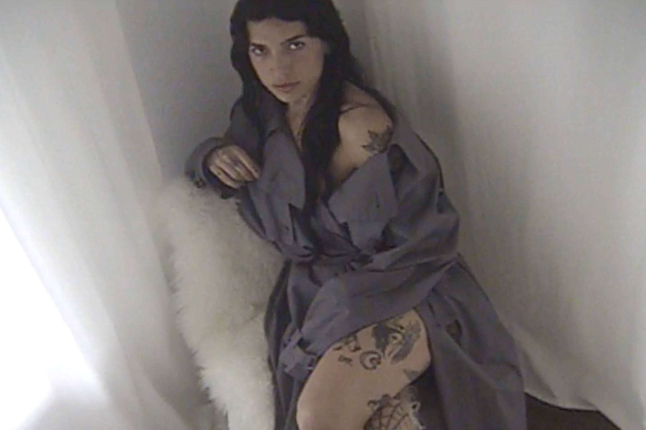 Woman with black hair and tattoos sitting on the floor in a small, enclosed space with white walls. She is wearing lace-up shoes, patterned tights, a jacket, and a top.