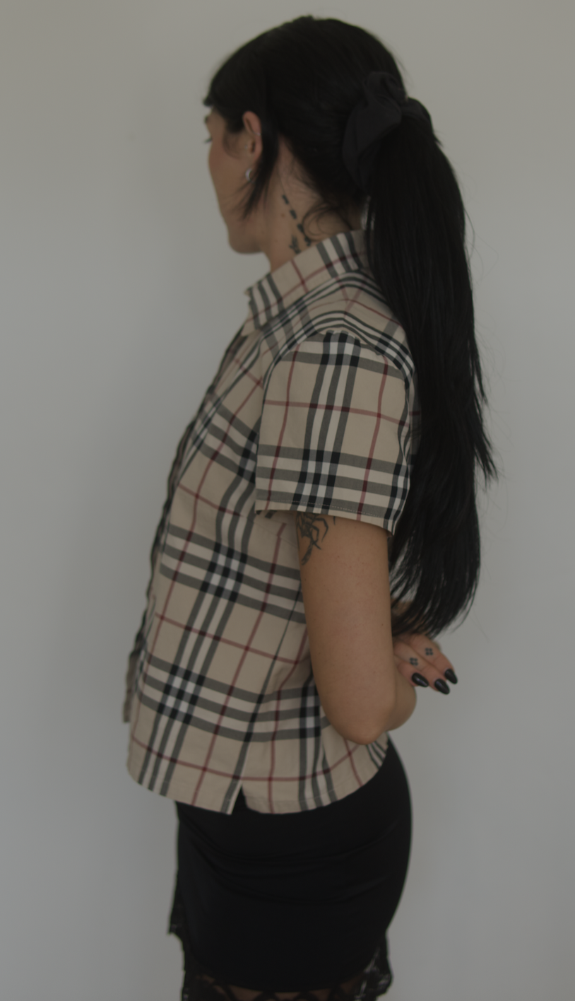 A woman with long black hair tied in a ponytail with a black ribbon, wearing a beige plaid shirt and black skirt, standing sideways against a plain white wall.