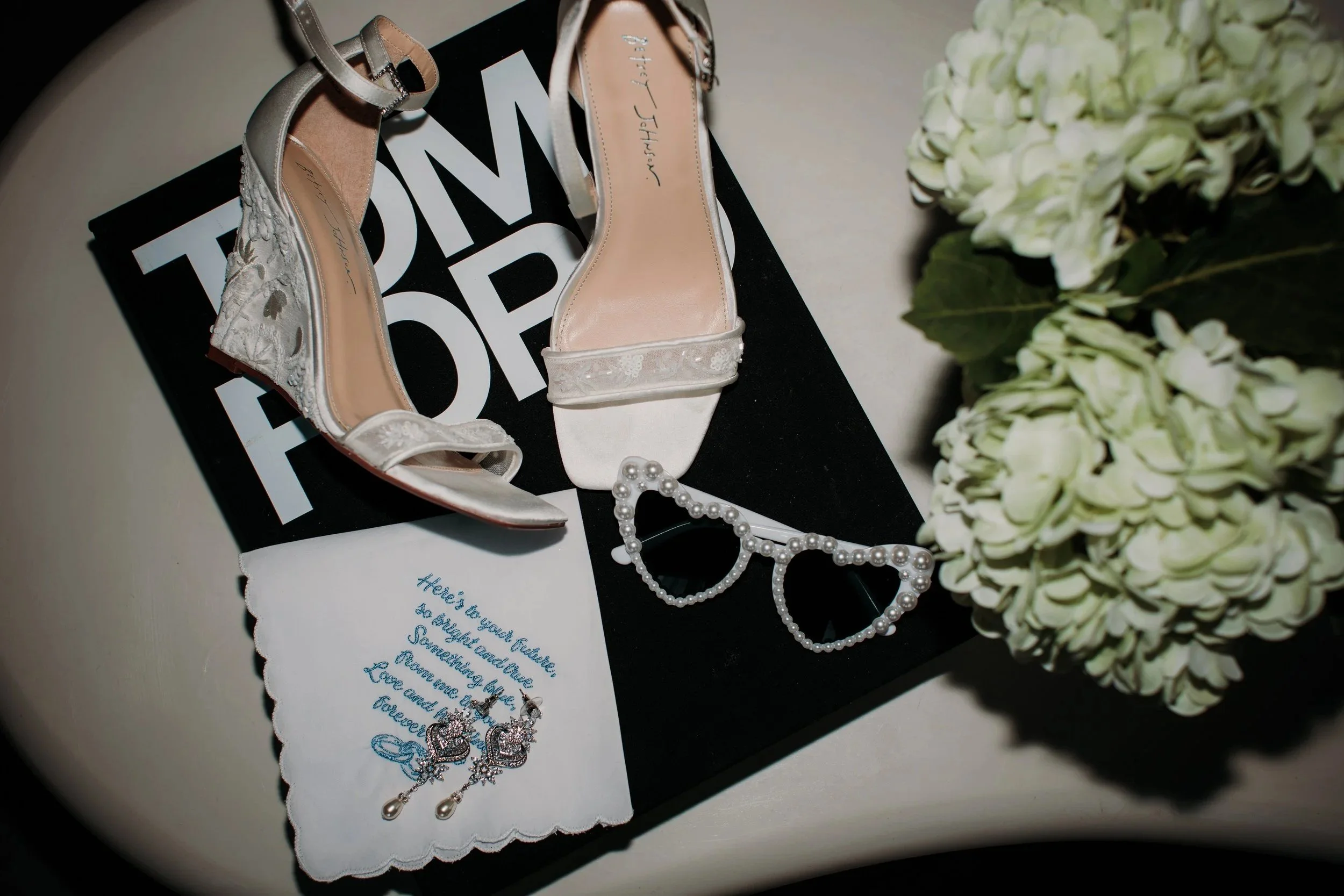 A pair of elegant white wedding shoes, a pearl bracelet and pearl earrings with a starfish design, sunglasses decorated with pearls, and a white handkerchief with blue embroidered text, all arranged on a black box with white lettering, next to a bouquet of white hydrangeas.