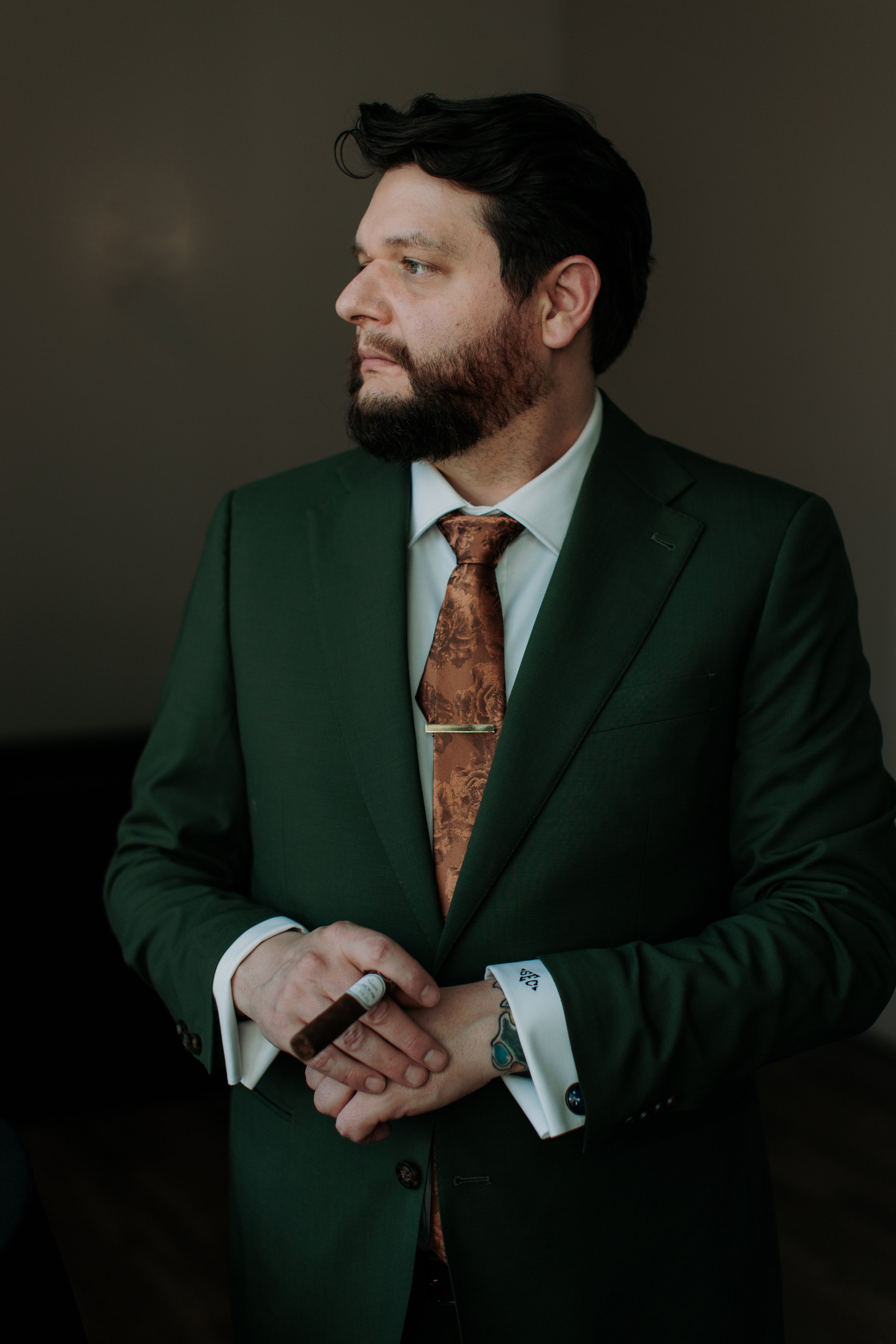 A man in a dark green suit, white shirt, and brown patterned tie holding a cigar, with tattoos visible on his hand and wrist, looking to the side.