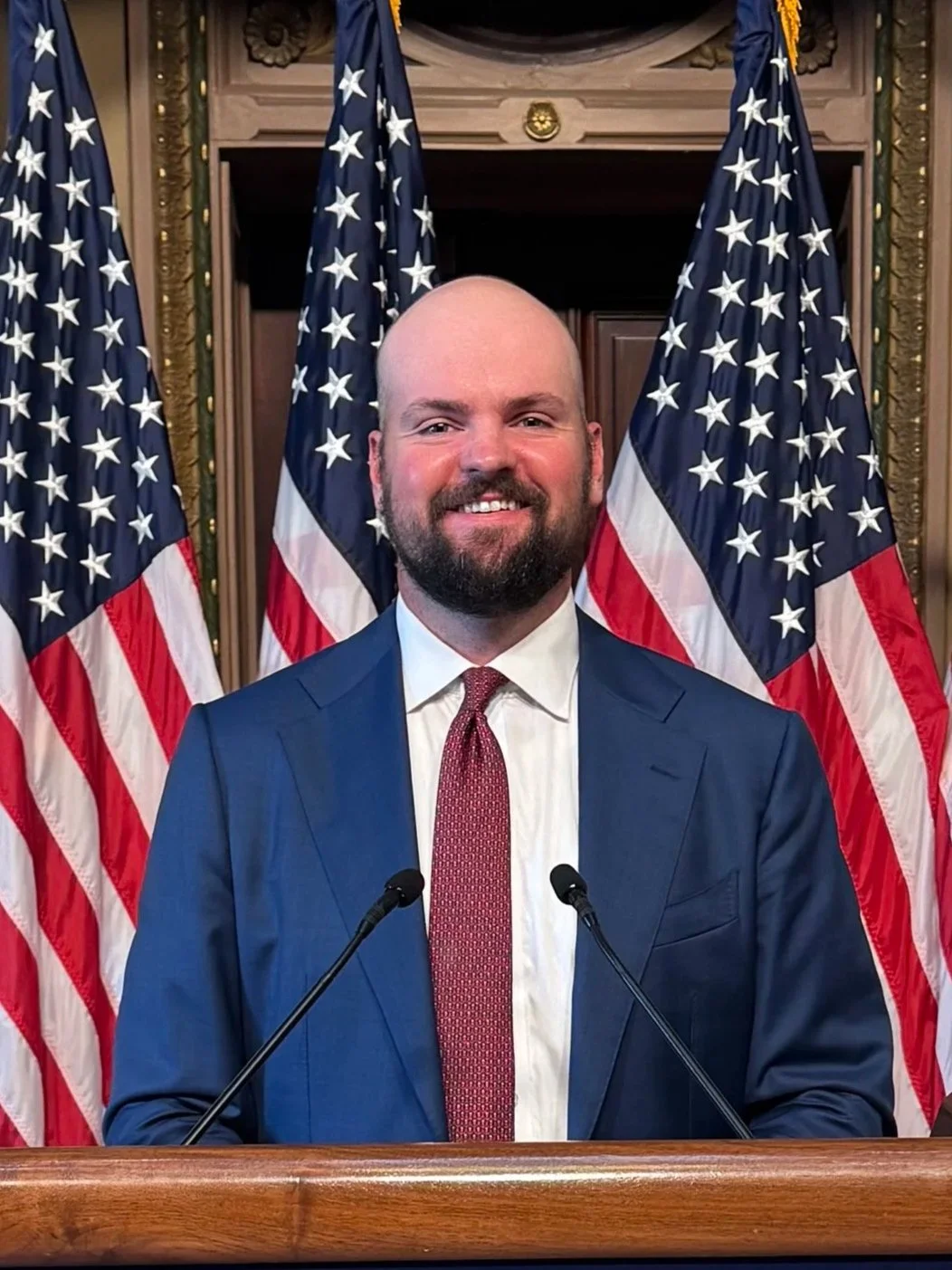 A man in a blue suit and red tie standing at a podium with microphones, smiling, with multiple American flags behind him in a formal setting.