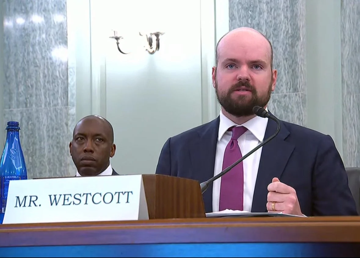 Man with a beard in a suit and purple tie speaking into a microphone at a hearing, with a nameplate reading 'Mr. Westcott' in front of him; another man in a suit is seated behind him.