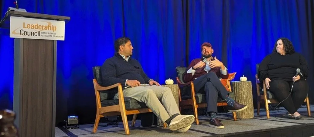 Three individuals seated on stage during a panel discussion at the Leadership Council, Small Business Association of Michigan, with a blue backdrop and pumpkin decorations.