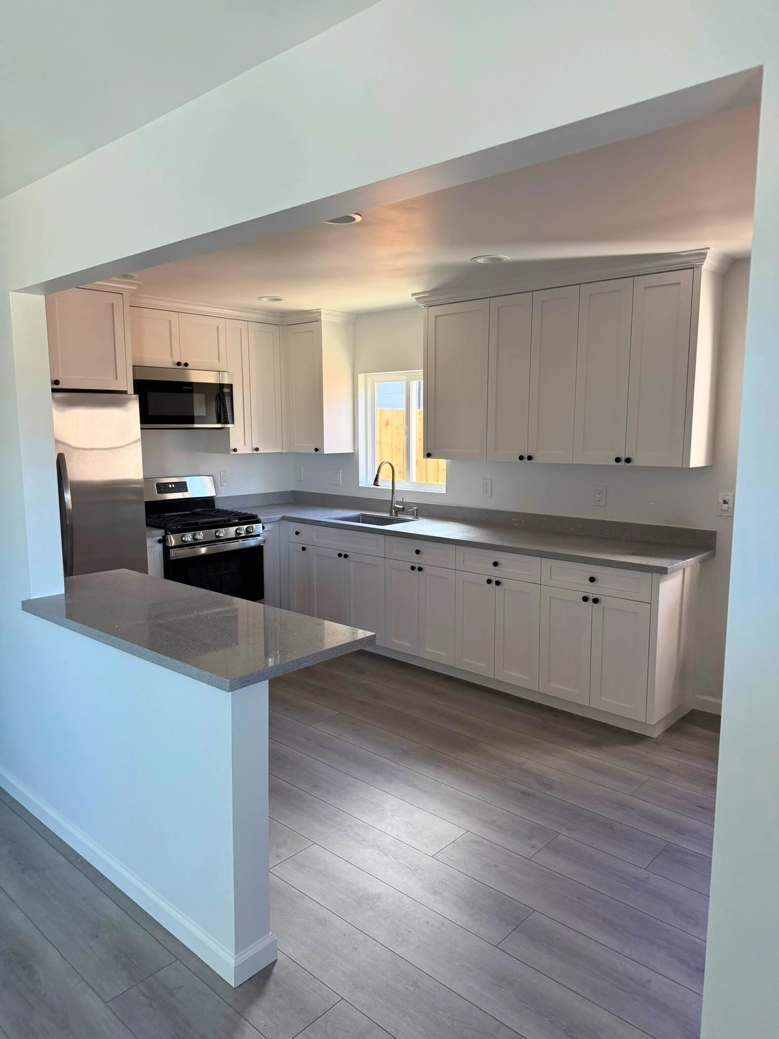Modern white kitchen with gray countertops and stainless steel appliances on hardwood floors, featuring upper and lower cabinets and a window over the sink.