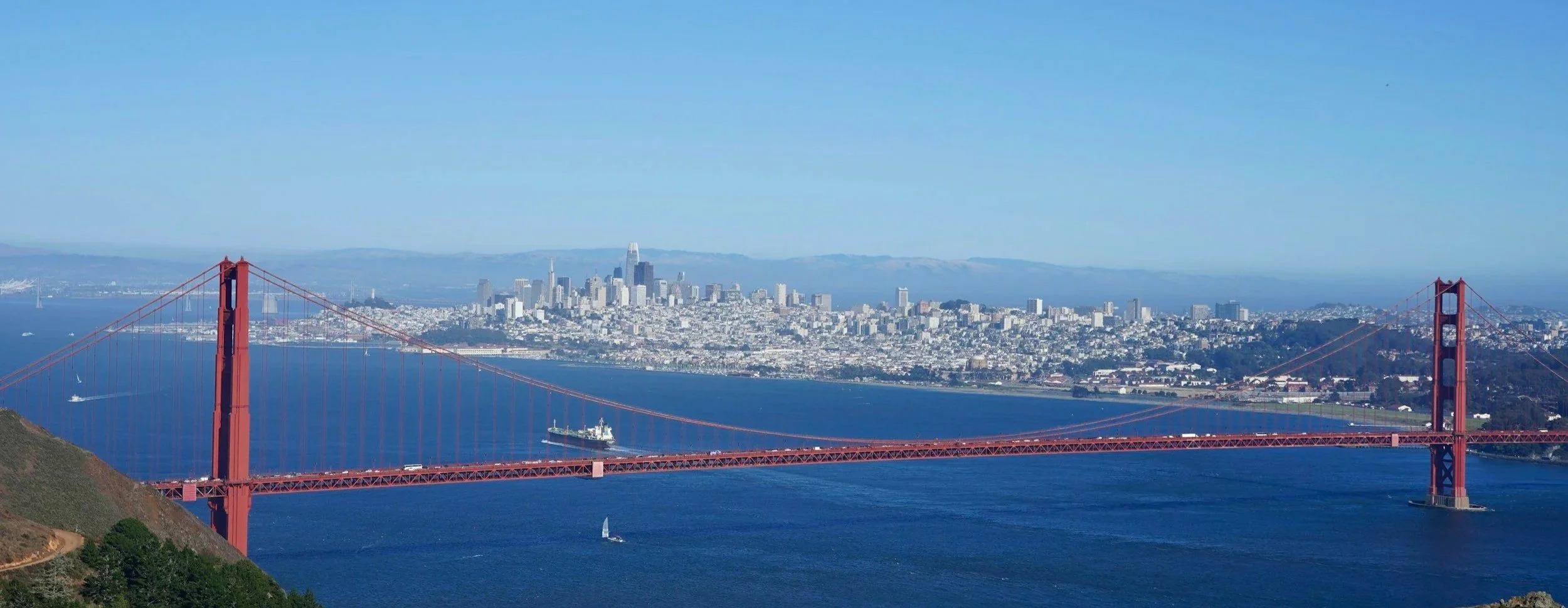 Panoramic view of the Golden Gate Bridge with San Francisco skyline in the background and boats in the water.