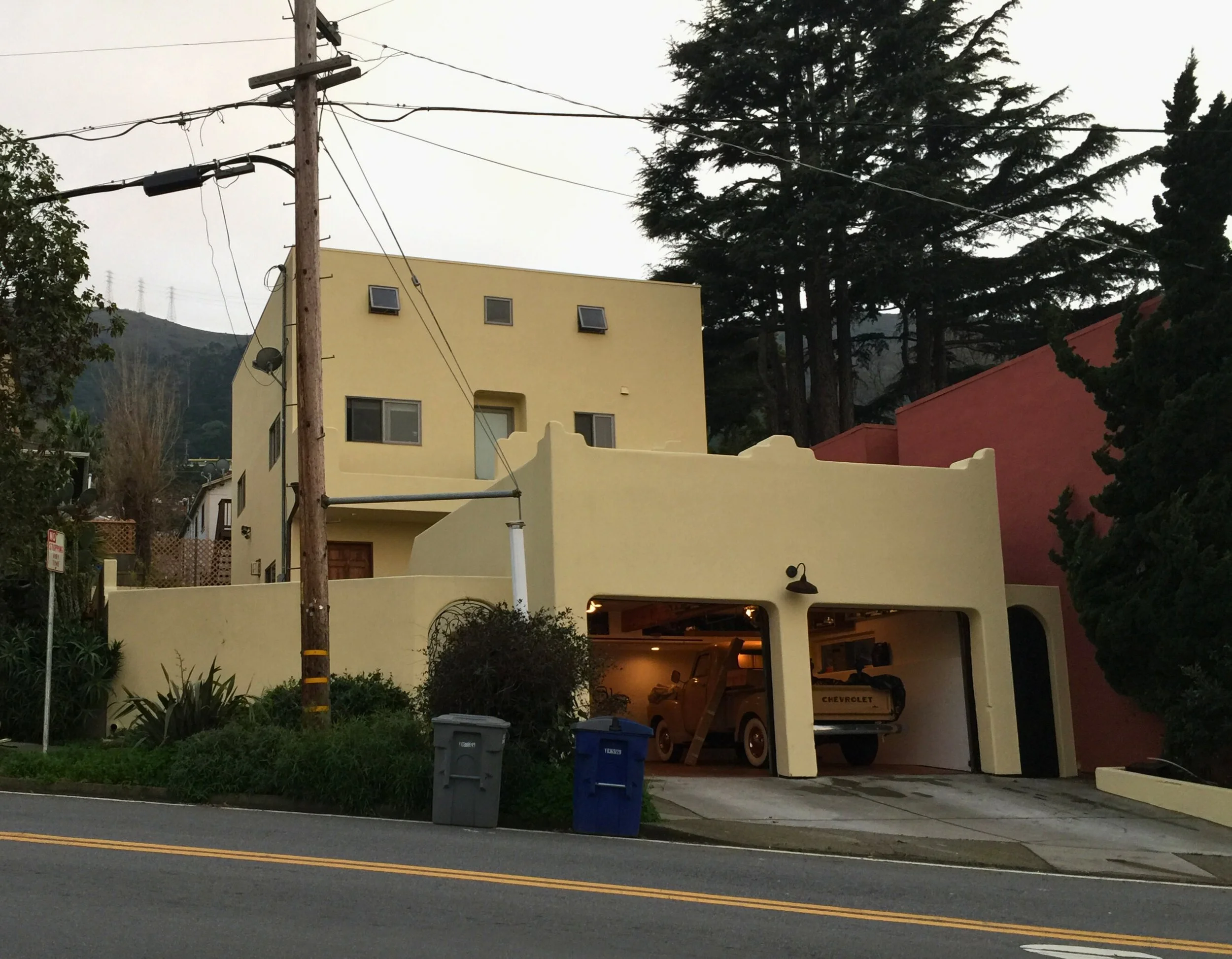 A multi-story house with a garage containing vintage vehicles, including a yellow Cheyenne pickup truck. The house has a yellow exterior, small windows, and a rounded prime area. Large trees are in the background, and there are utility poles and street signs in the foreground.