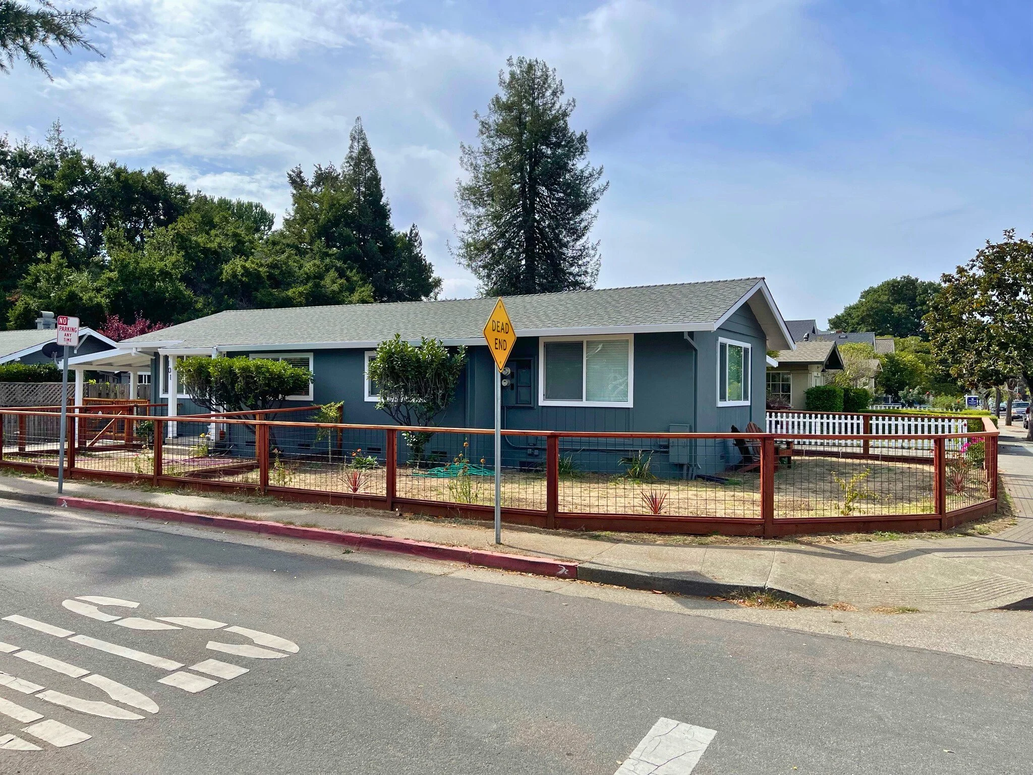 A small blue house with white trim and a gray roof, surrounded by a red fence. There are two trees in the front yard, a ‘Dead End’ sign in front of the house, and a 'No Parking' sign on the corner near the street. Sidewalks and a crosswalk are visible in the foreground.
