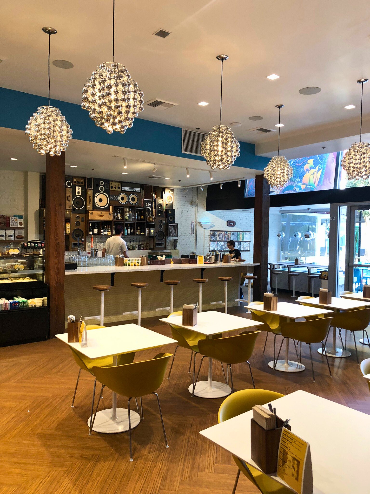 Interior of a modern cafe with multiple tables and yellow chairs, a barista counter with bar stools, hanging chandeliers, and a bright display screen near the entrance.