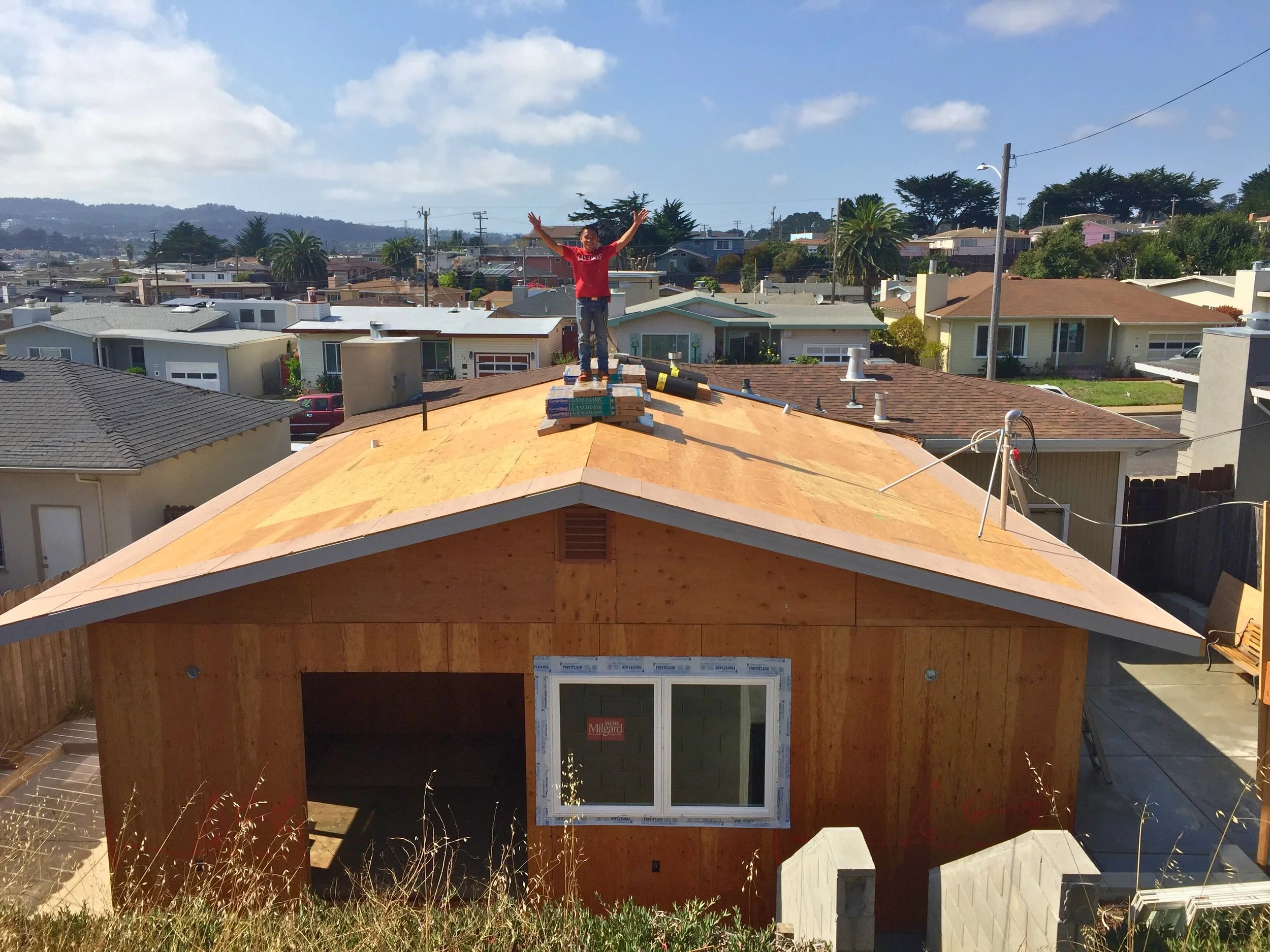 A person standing on top of a house under construction with arms raised, surrounded by wooden framing and insulation material, in a neighborhood with other houses and trees in the background.
