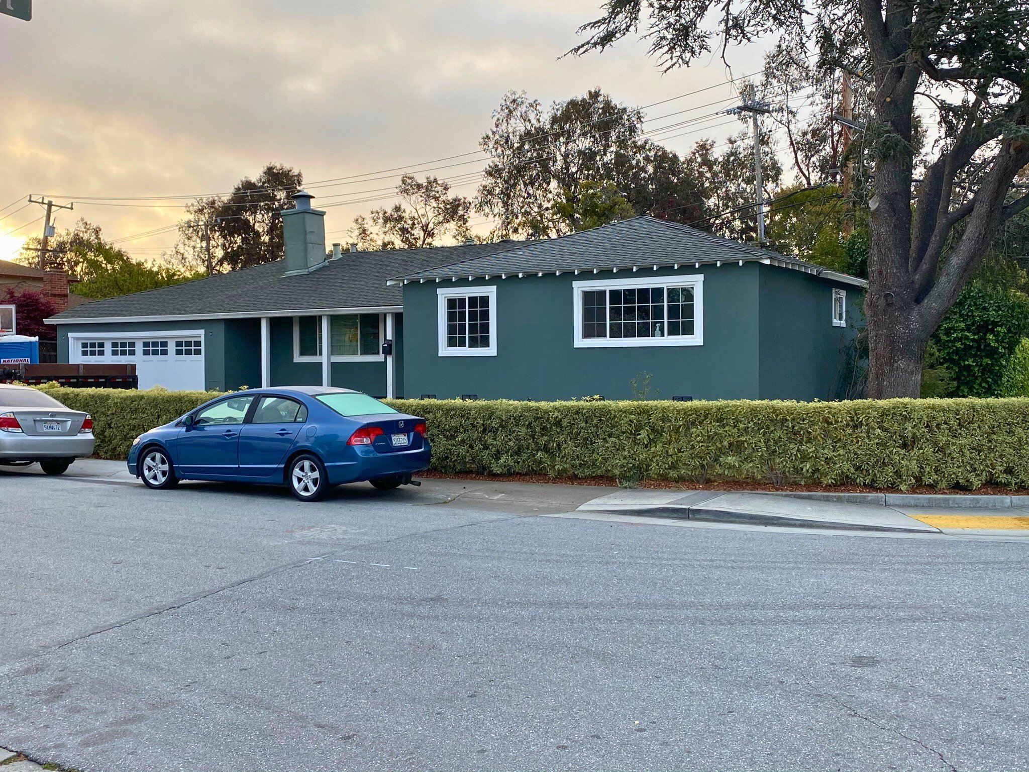 A green house with white trim and shingle roof, with two cars parked in front on a street, and a large tree to the right. The background has a cloudy sky and utility poles.