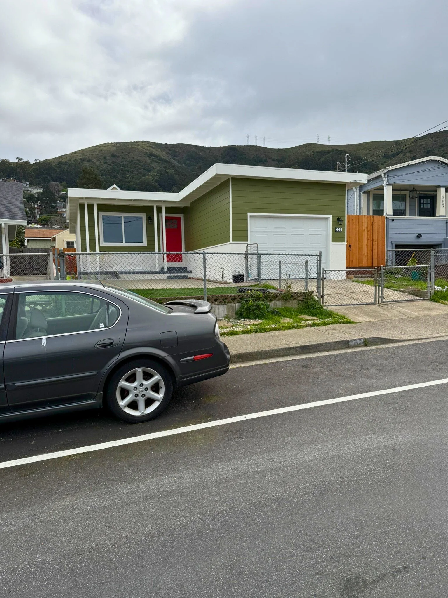 A green, modern single-story house with a white garage door and red front door, situated behind a chain-link fence, with a black car parked on the street in front, and a mountain landscape with cloudy sky in the background.