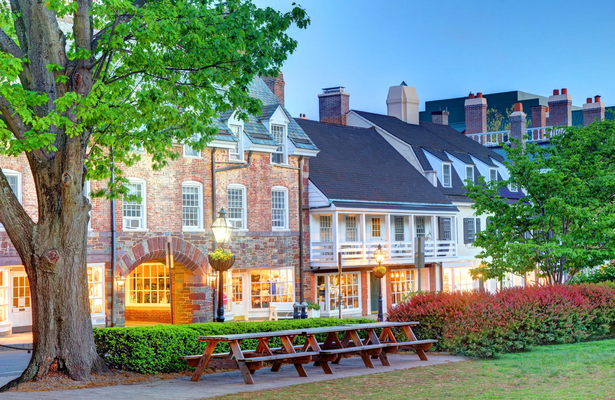 A street view of a row of brick buildings with white and gray roofs in Palmer Square in Princeton, NJ , illuminated by warm outdoor lighting, with trees and bushes in the foreground and a clear blue sky in the background.
