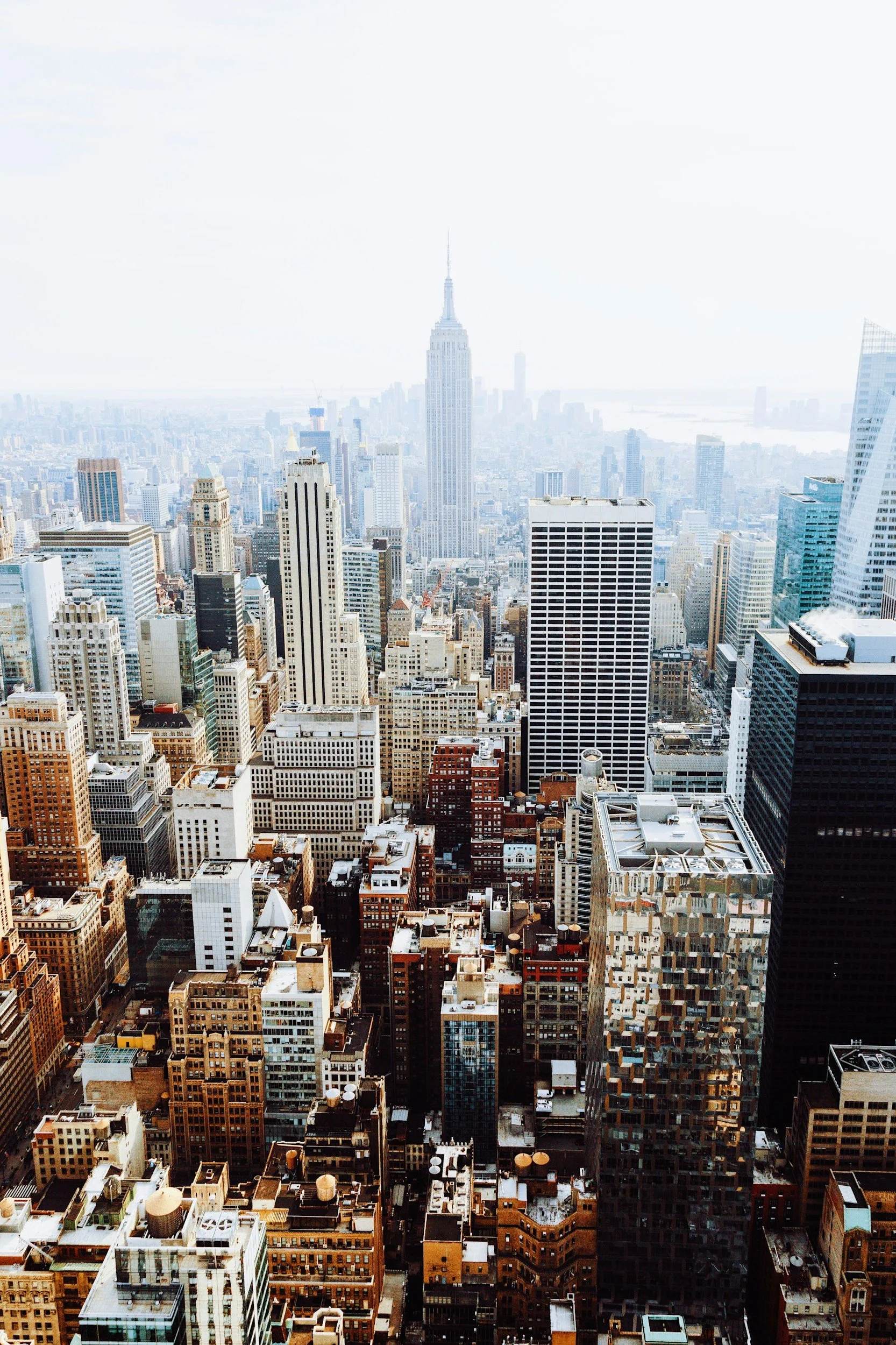New York skyline featuring tall buildings, with the Empire State Building prominently in the center. The photo appears to be taken from a high vantage point, showing other skyscrapers in the foreground and background, with a bright sky overhead.