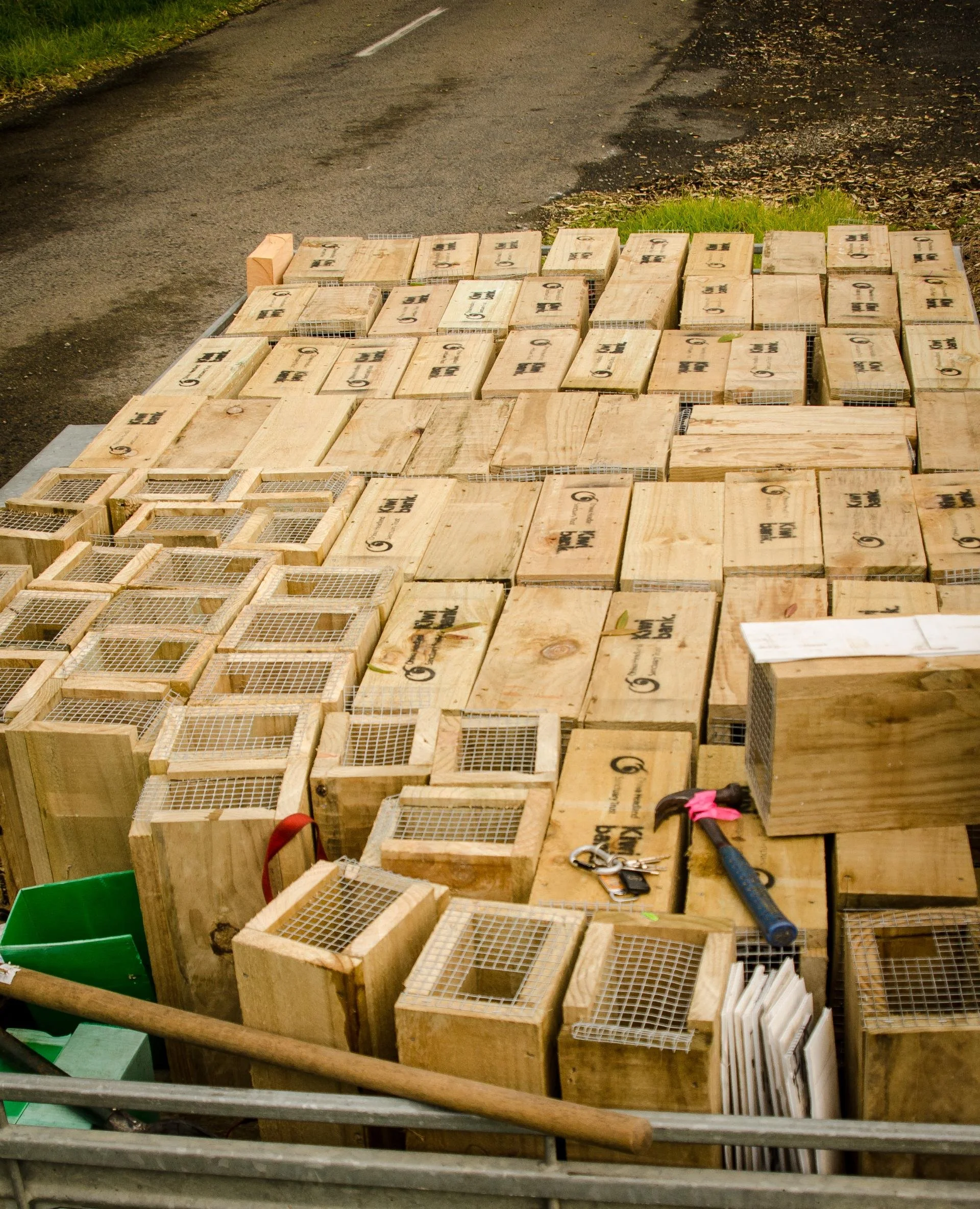 Stacked wooden boxes with metal wire mesh tops, arranged on the ground alongside a gravel road, with tools and papers nearby.