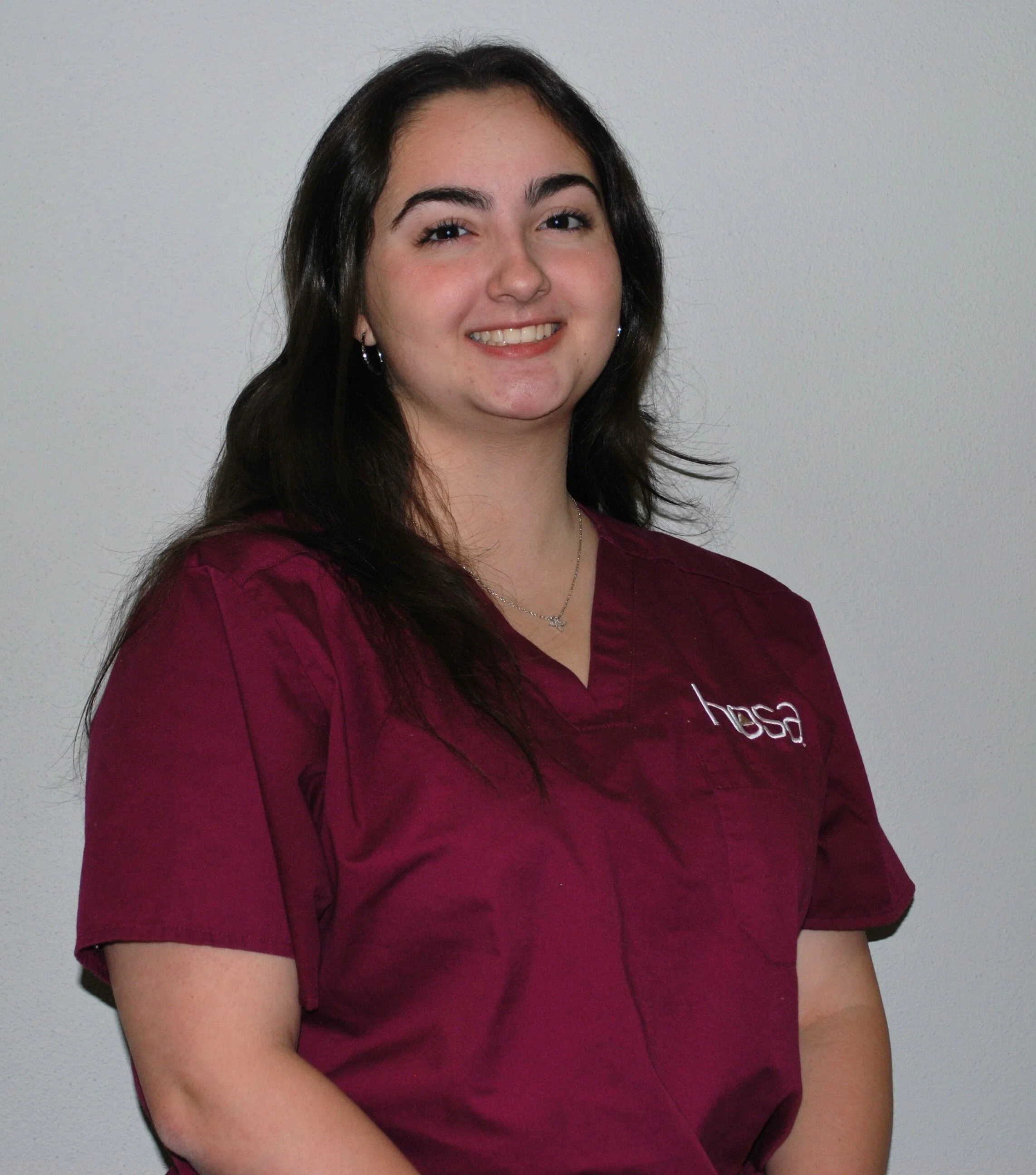 A young woman with long dark hair and earrings, wearing a maroon medical uniform with 'hosa' embroidered on it, standing against a plain white wall.