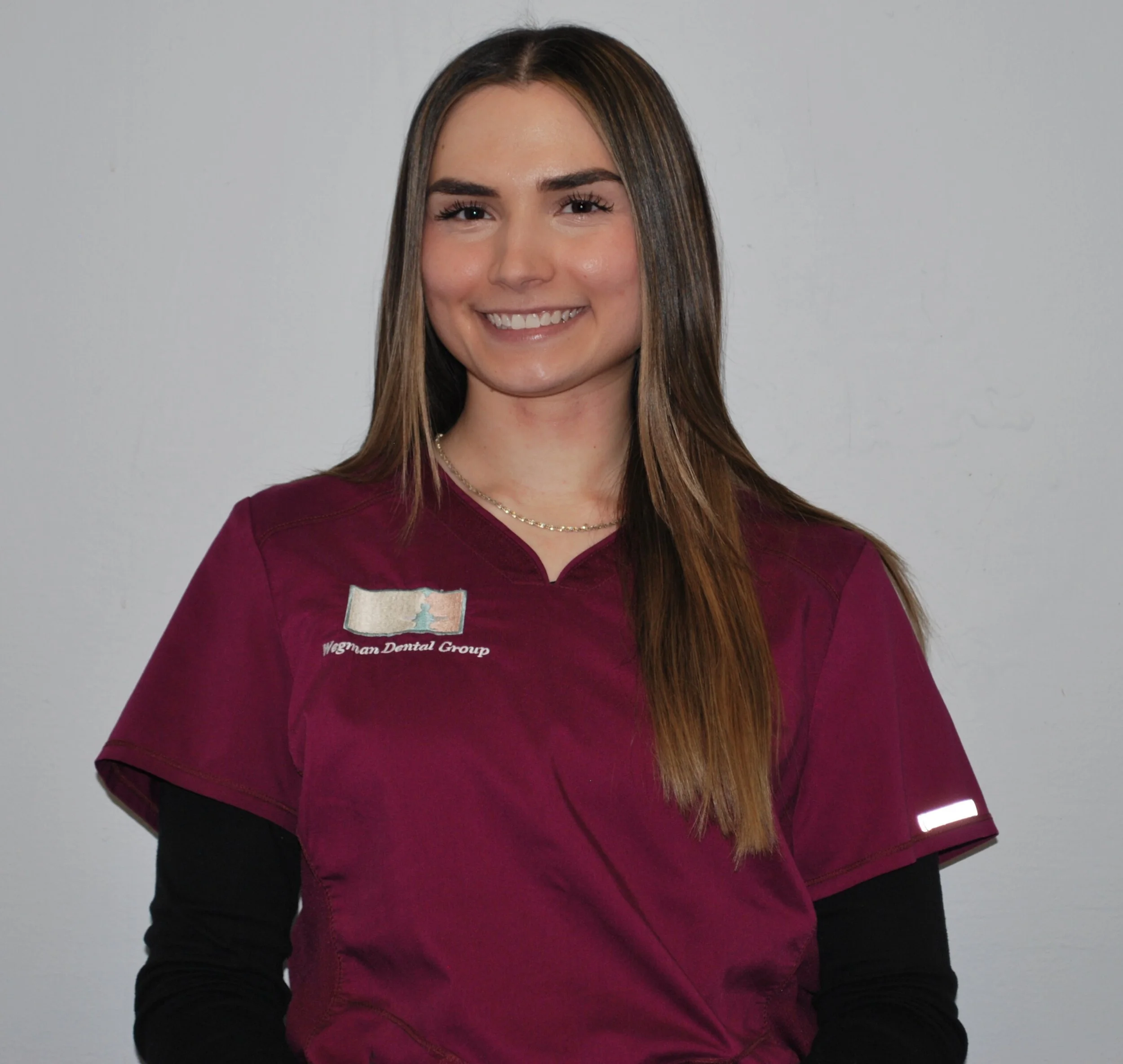 Smiling woman with long brown hair wearing dental scrubs with a logo that reads 'Wogman Dental Group' standing against a plain gray background.