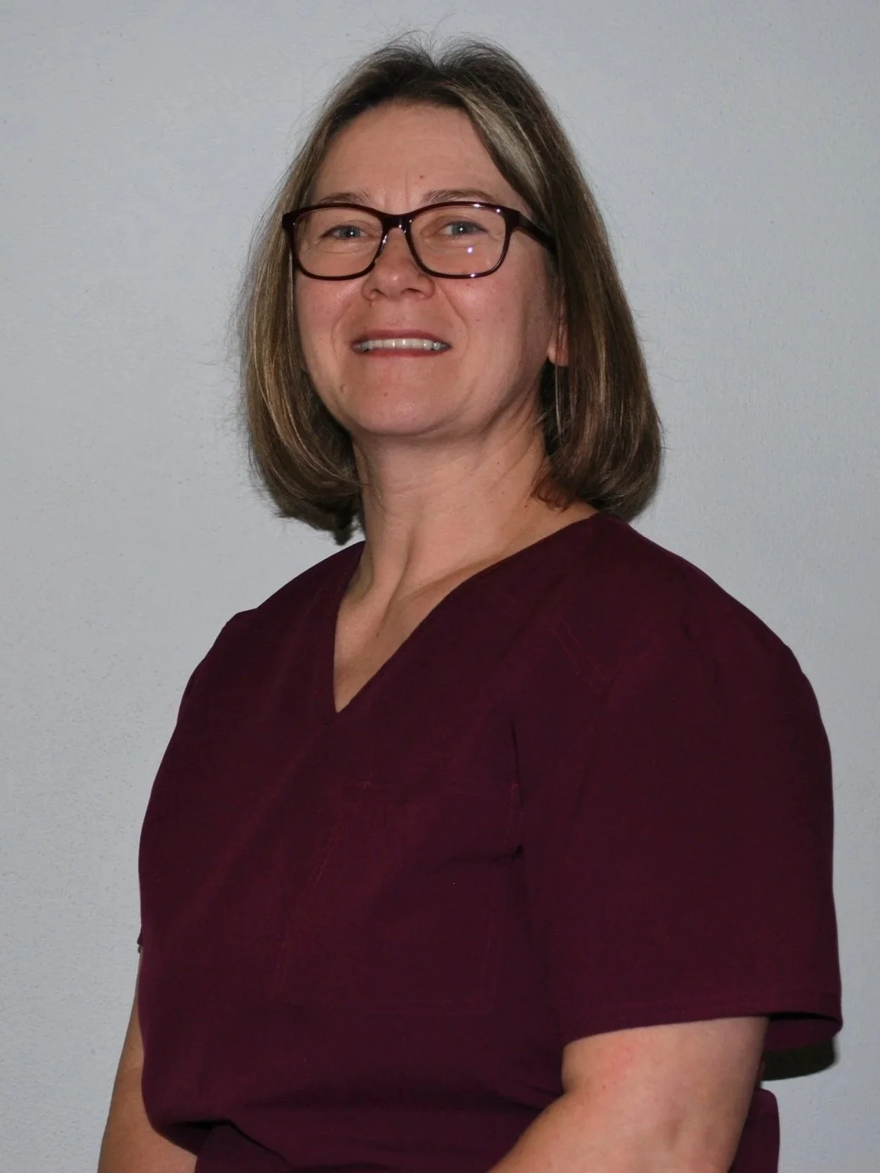 A woman with shoulder-length brown hair, glasses, and a maroon shirt standing against a plain light gray wall.