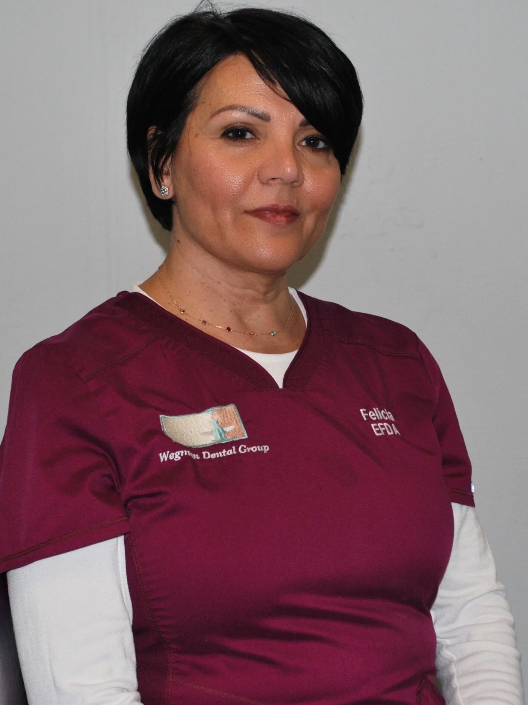 A woman with short black hair wearing maroon medical scrubs with 'Wegmans Dental Group' and her name 'Felicity' embroidered on them, standing against a plain white wall.