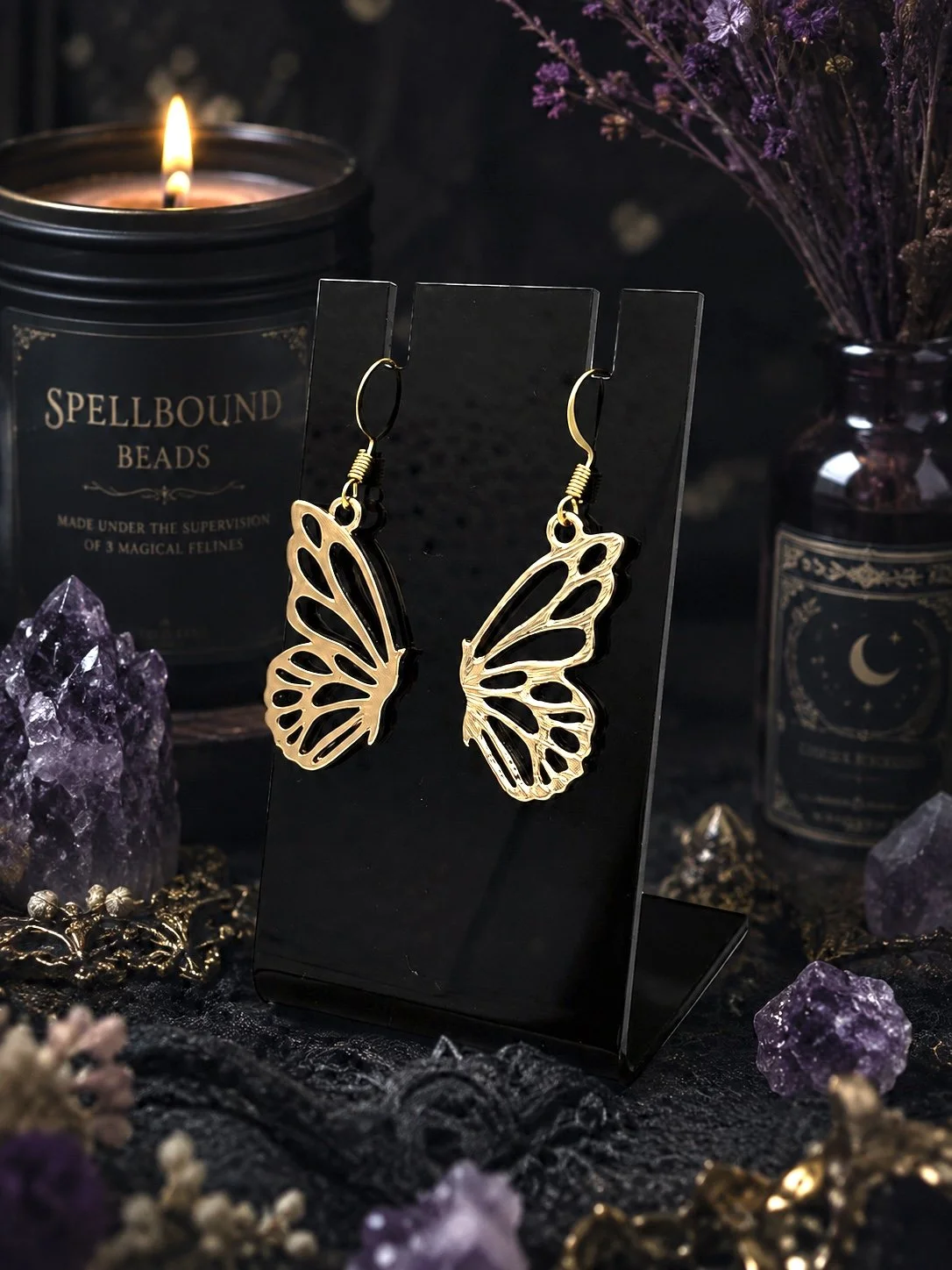 Gold butterfly-shaped earrings displayed on a black stand, surrounded by amethyst crystals, dried flowers, and candles in dark containers with mystical designs.